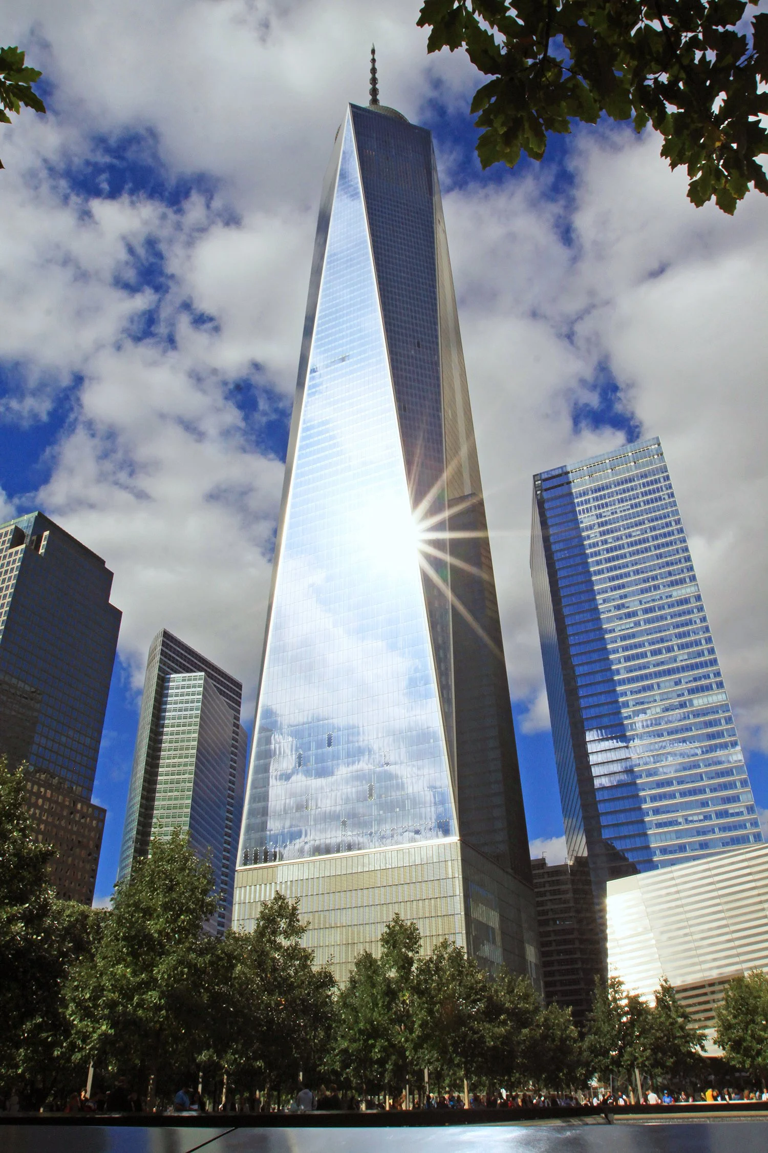 A tall, glass skyscraper in New York City, with a reflection of the sky and clouds, surrounded by other high-rise buildings and trees. Freedom Tower.