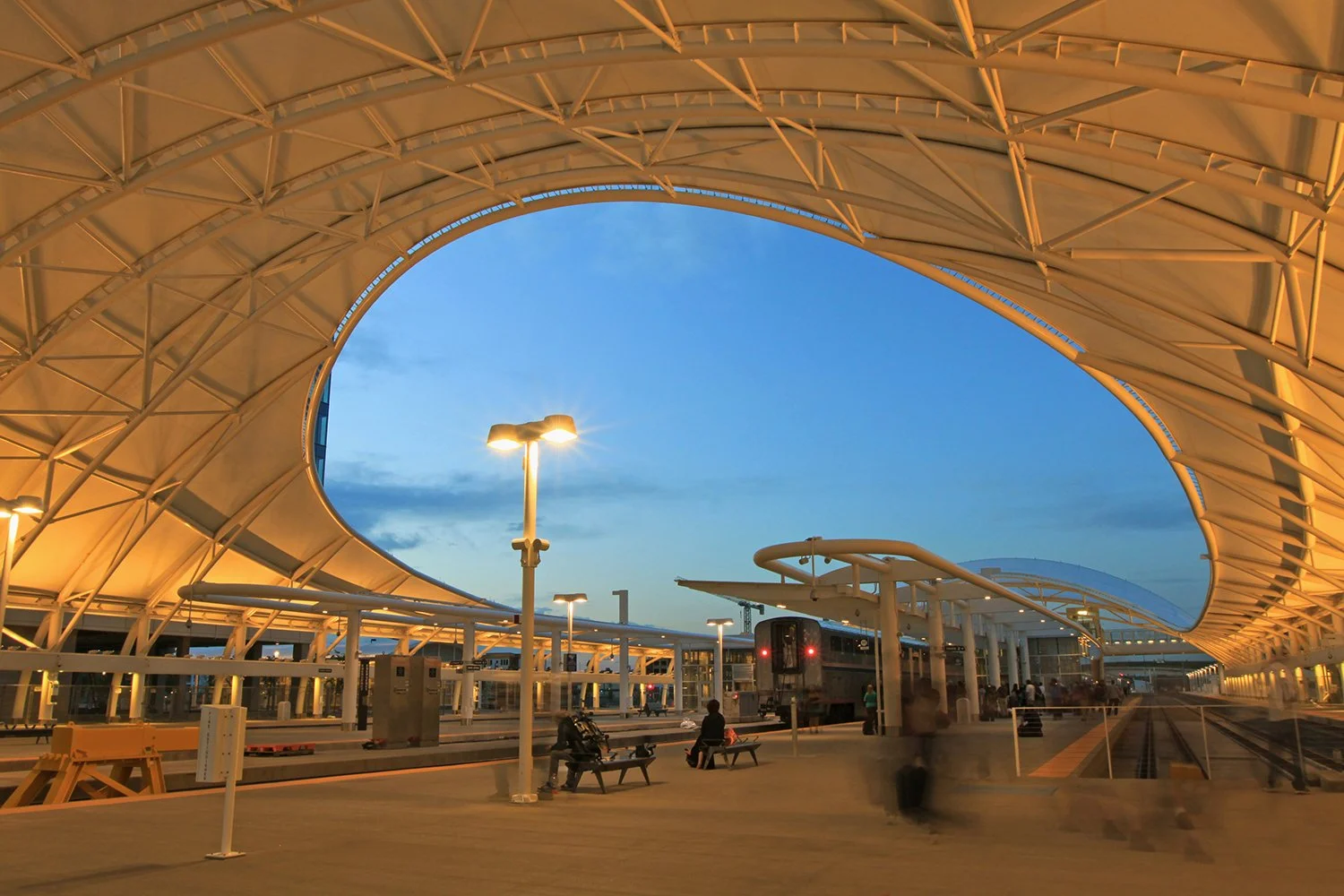 Denver train station with curved white roof during evening, with waiting passengers, benches, and train tracks.