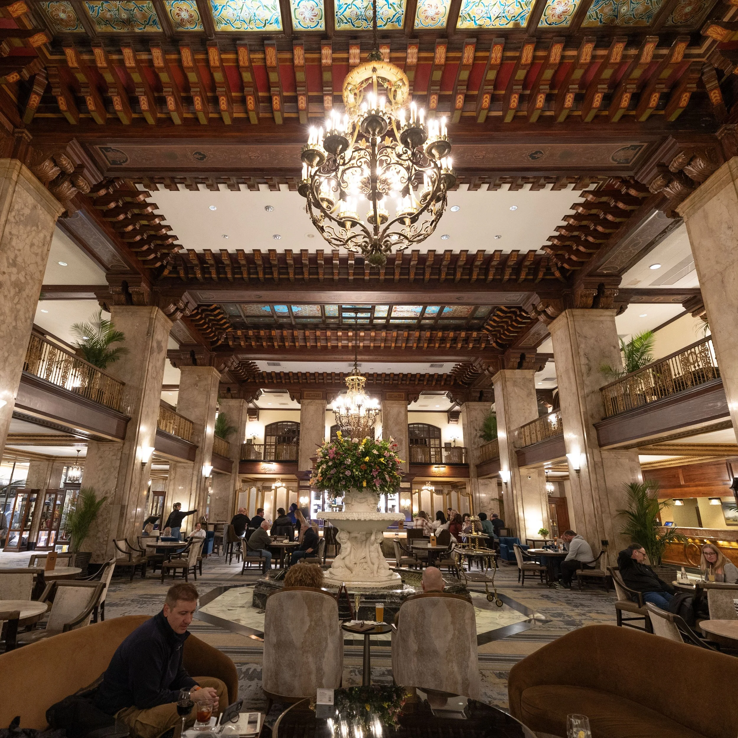A grand hotel lobby with marble columns, a large floral arrangement on a fountain, and ornate chandeliers hanging from a decorated wooden ceiling. Peabody Hotel Memphis.