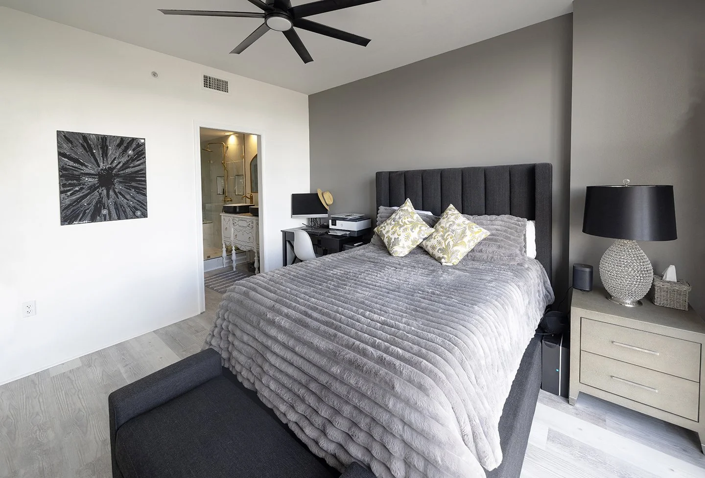 A modern bedroom with a gray upholstered bed, decorative pillows, a black nightstand with a lamp, and a ceiling fan. High-rise condominium in Atlanta.