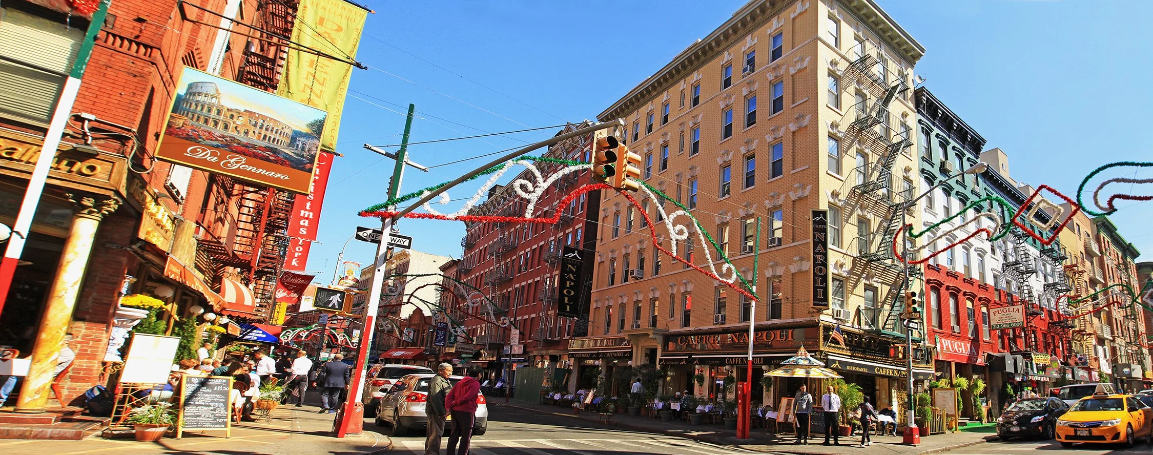 Street scene in New York City with colorful buildings, overhead holiday lights, pedestrians, parked cars, and a restaurant named 'Caffe Napoli'. Little Italy.