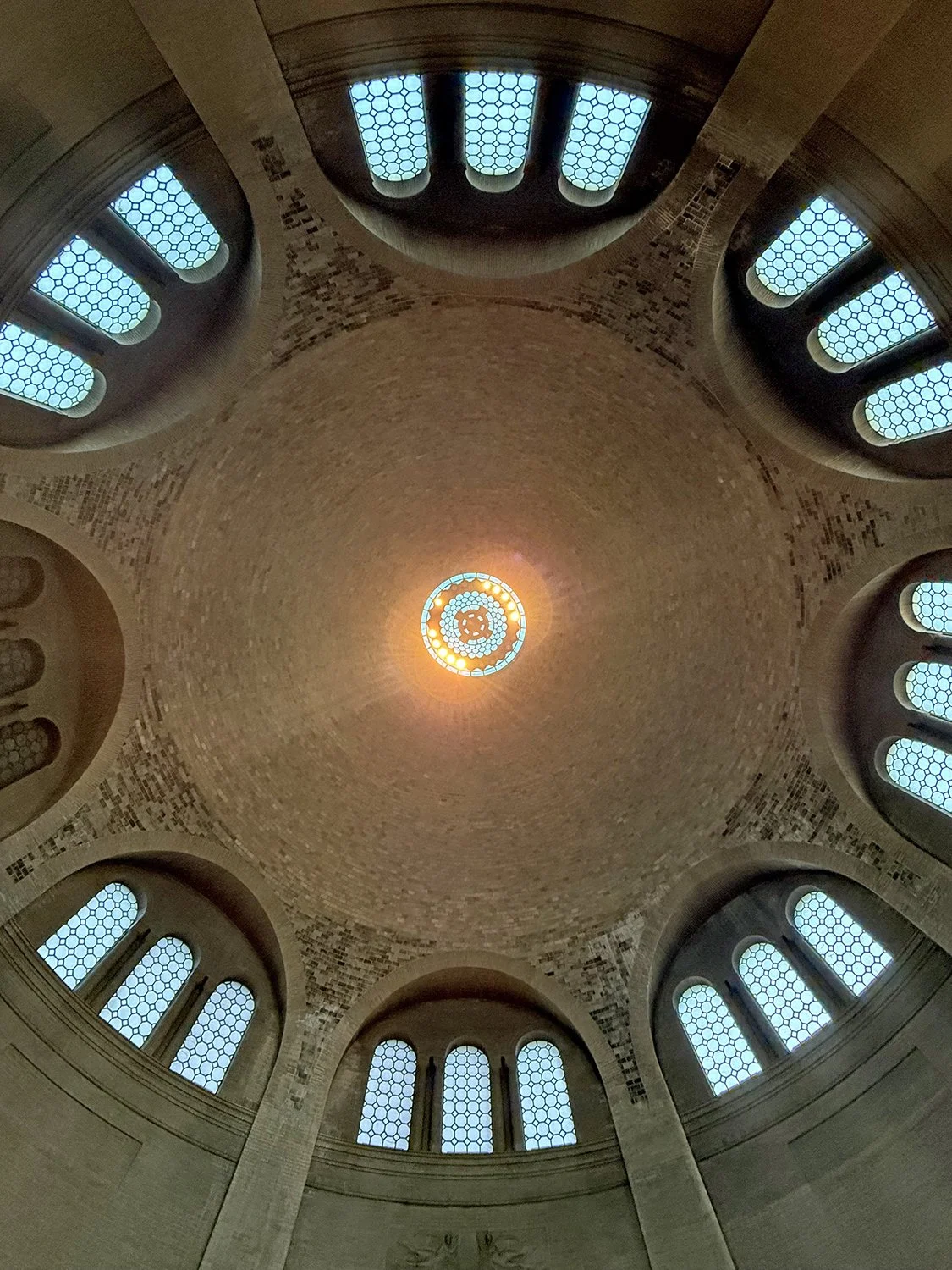 Looking up at the interior of a domed ceiling with skylights, featuring a circular light fixture at the center, and arched windows around the perimeter with hexagonal patterned glass. Penn Museum Philadelphia.