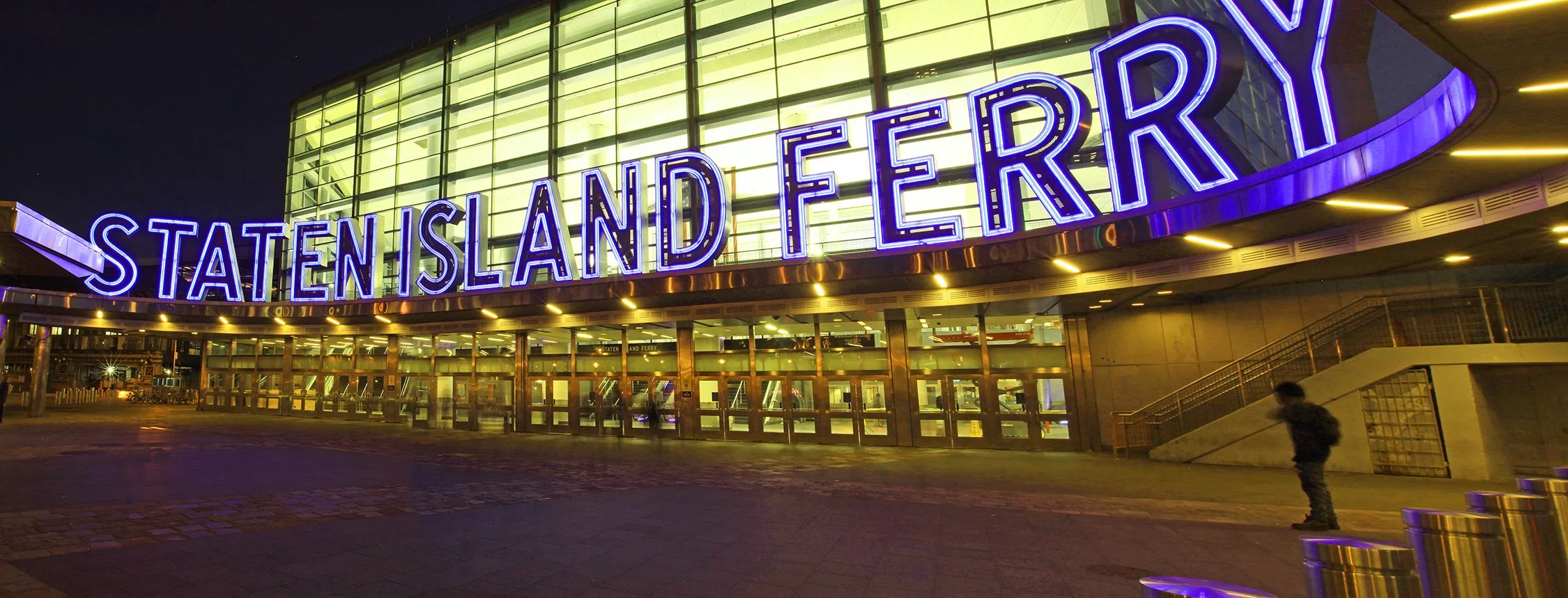 Night view of the Staten Island Ferry terminal with illuminated sign reading 'STATEN ISLAND FERRY' in purple neon lights and glass facade.