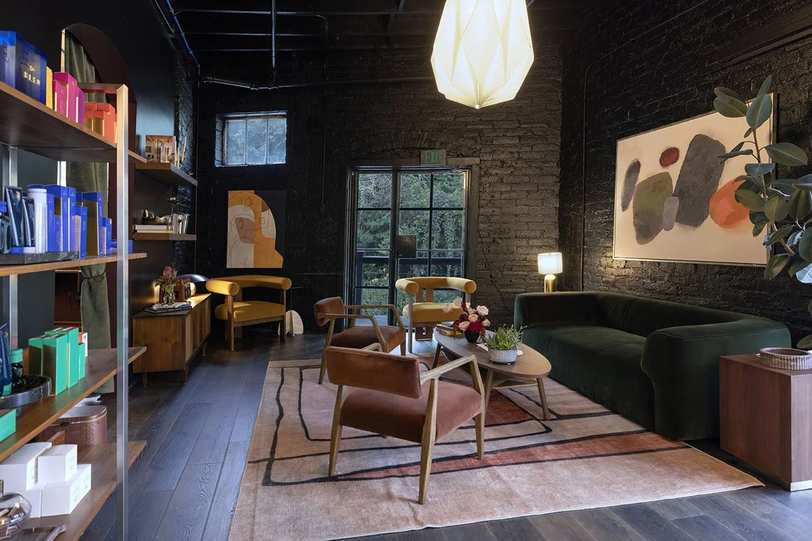 A cozy modern lounge with black brick walls, a large window, and a white pendant light hanging from the ceiling. The room features a green velvet sofa, yellow armchairs, a round coffee table with flowers, and abstract art on the wall. Le Babe Atlanta