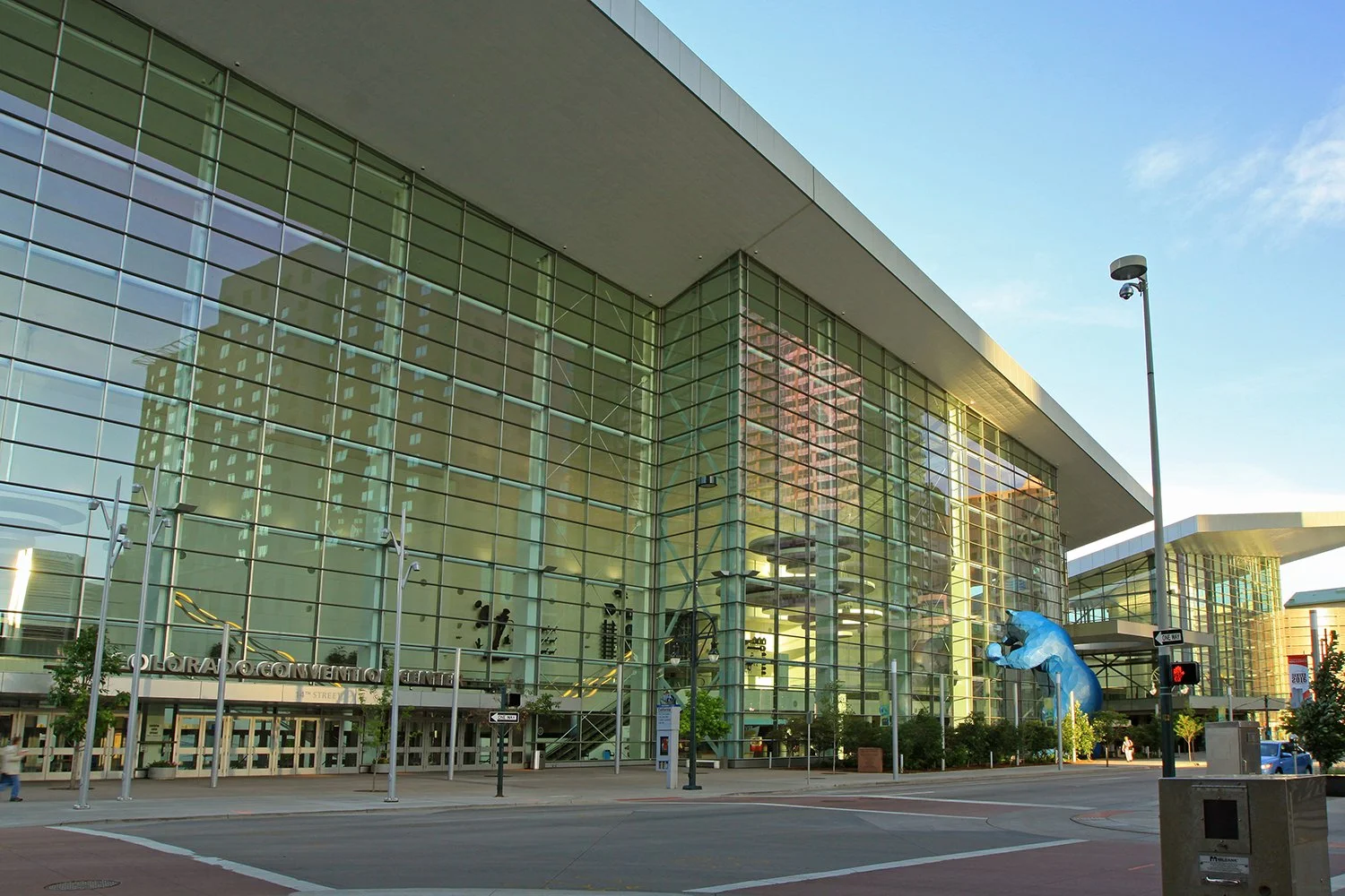 Exterior view of Denver Convention Center. There are trees, streetlights, and a large blue bear sculpture outside. Daytime.