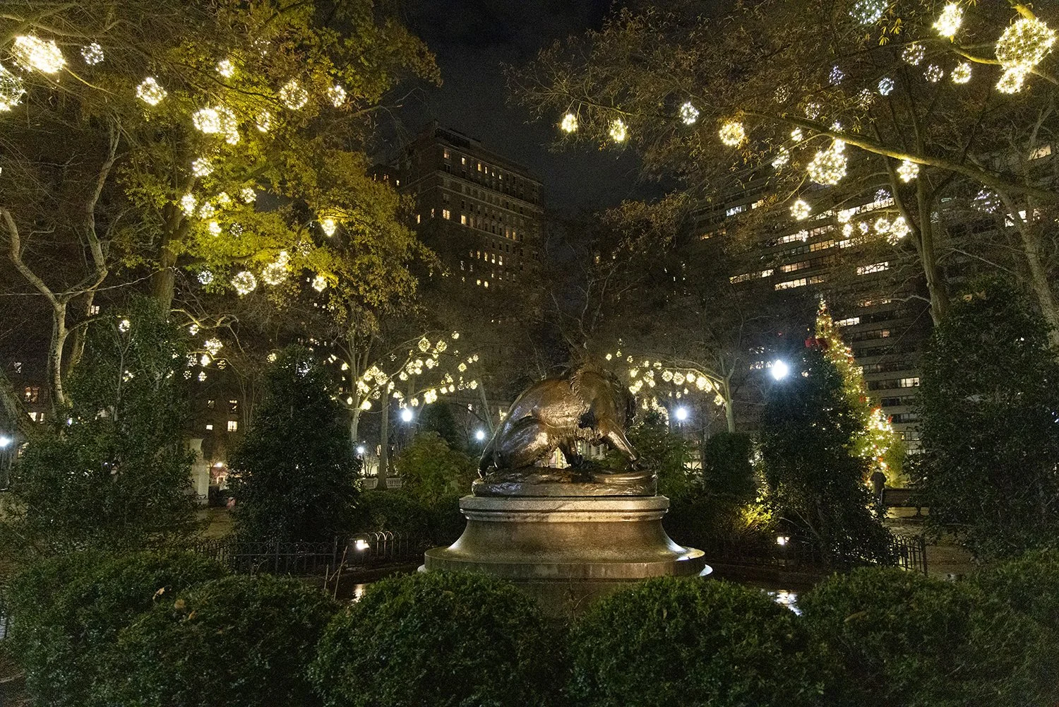 A night view of a park with a statue of a rat on a pedestal, surrounded by trees decorated with hanging string lights and a Christmas tree with lights in the background.