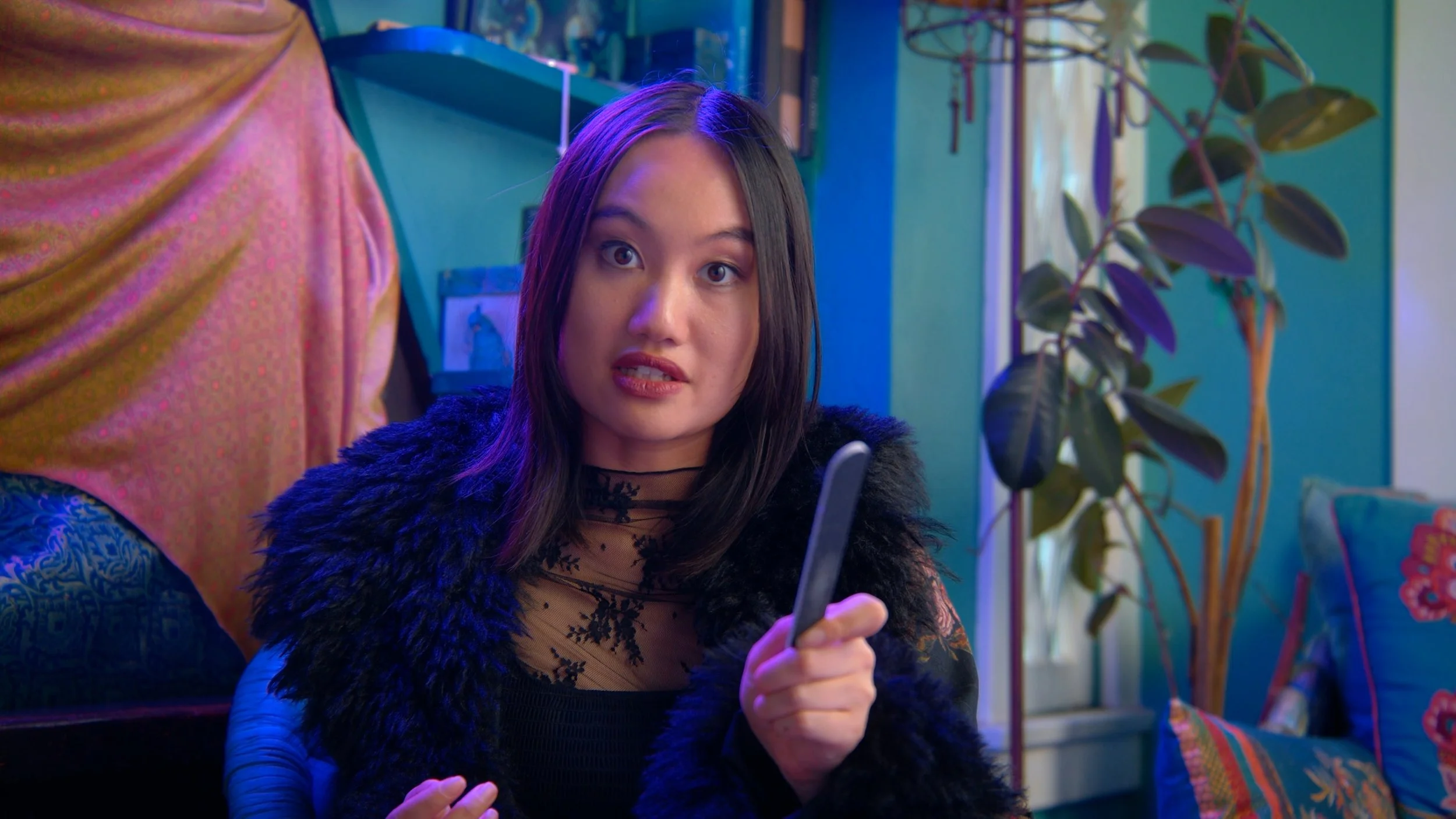 A young woman with shoulder-length dark hair holding a knife in her right hand, sitting in a colorful room with patterned pillows, a large potted plant, and vibrant lighting.