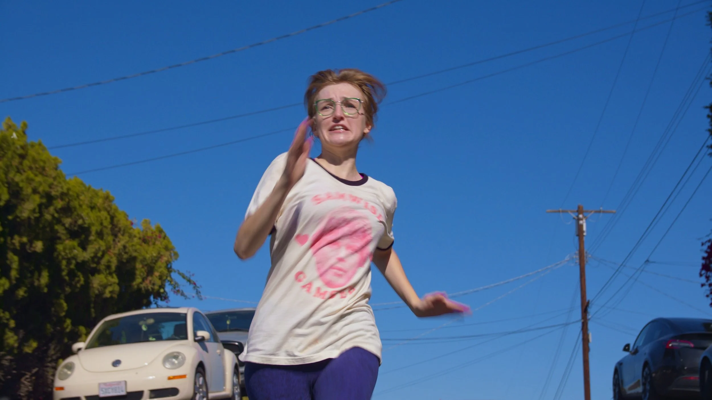 A woman with glasses and short hair running outside on a sunny day, wearing a white T-shirt with a pink graphic of a face and the words "Sean Spicer A Meme." There are cars and trees in the background.