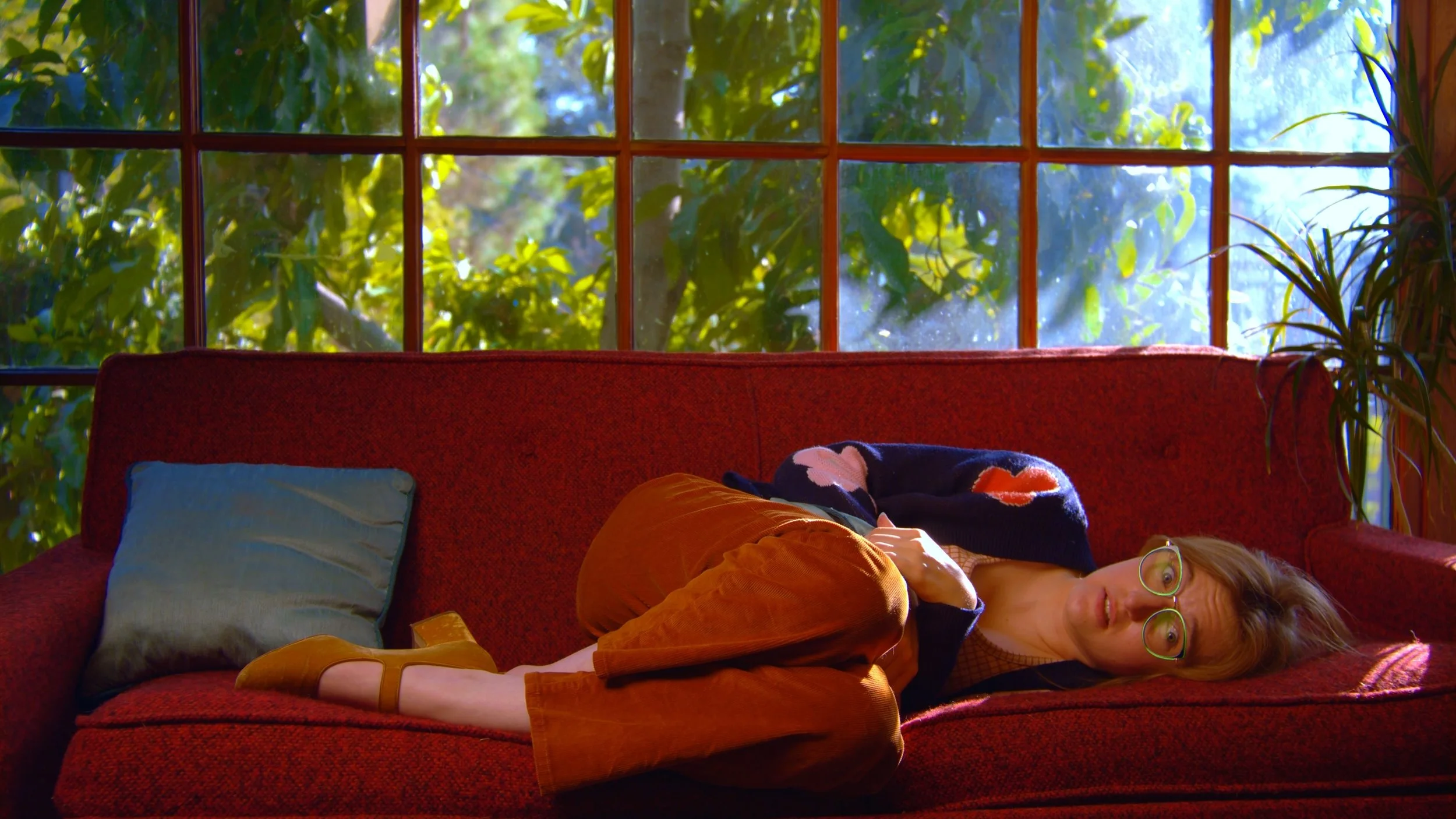 Woman lying on a red couch with a greenish pillow near the window with green foliage outside. She has glasses, reddish hair, and is wearing brown pants and a dark sweater.