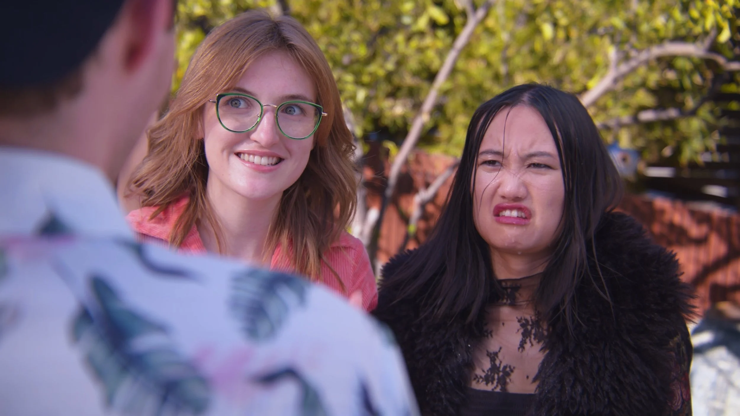 Two women standing outside in front of trees, one smiling with glasses and the other making a disgusted face.