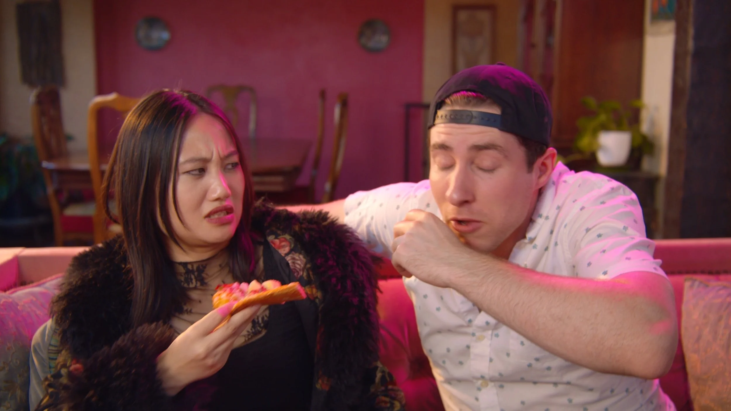 A woman and a man sitting on a pink couch in a room with pink and beige walls. The woman is holding a slice of pizza and looking confused, while the man is sneezing or coughing.