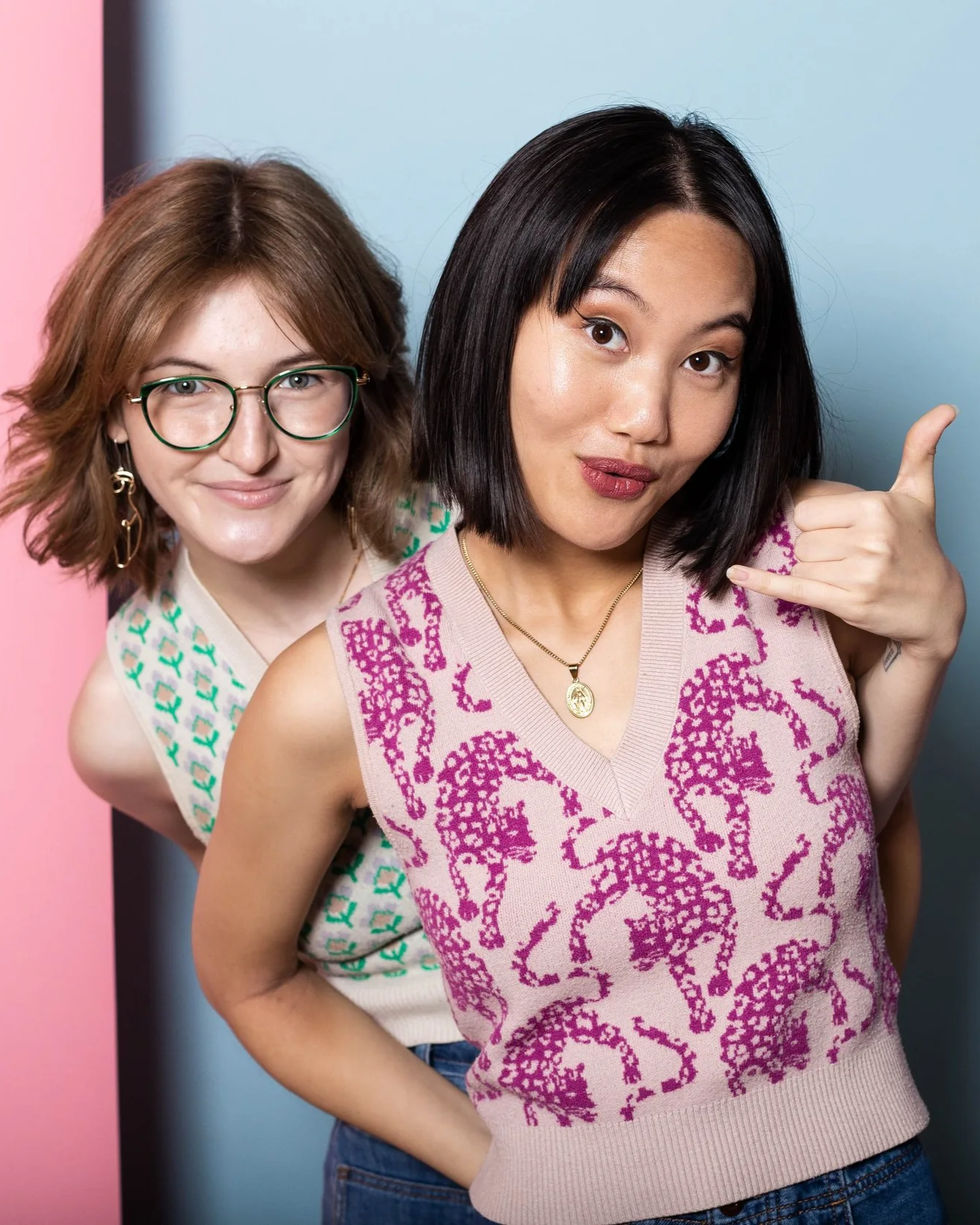 Two women posing together, one making a call me gesture with her hand, smiling, in front of colorful pink and blue background.