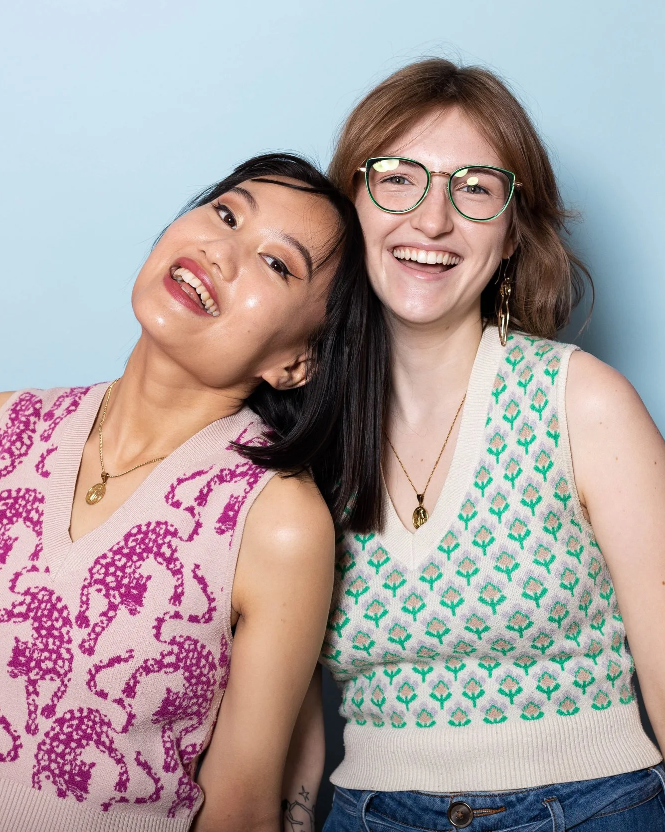 Two women smiling and leaning on each other against a light blue background, one with Asian features and black hair, the other with light skin, red hair, and glasses.