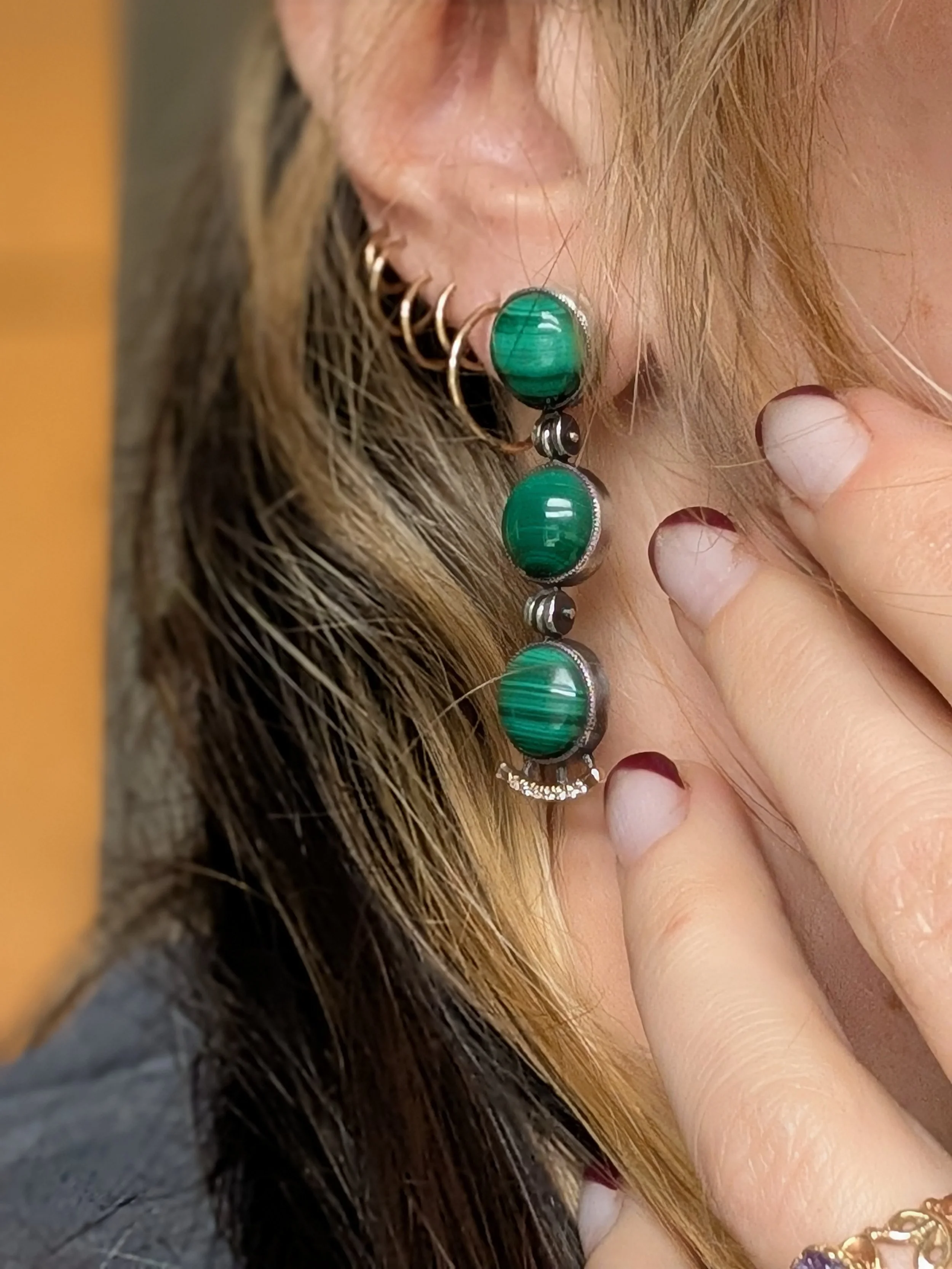 Close-up of a person's ear adorned with multiple earrings, including a large, oval, green gemstone earring and several gold hoops, with a hand touching the earlobe.