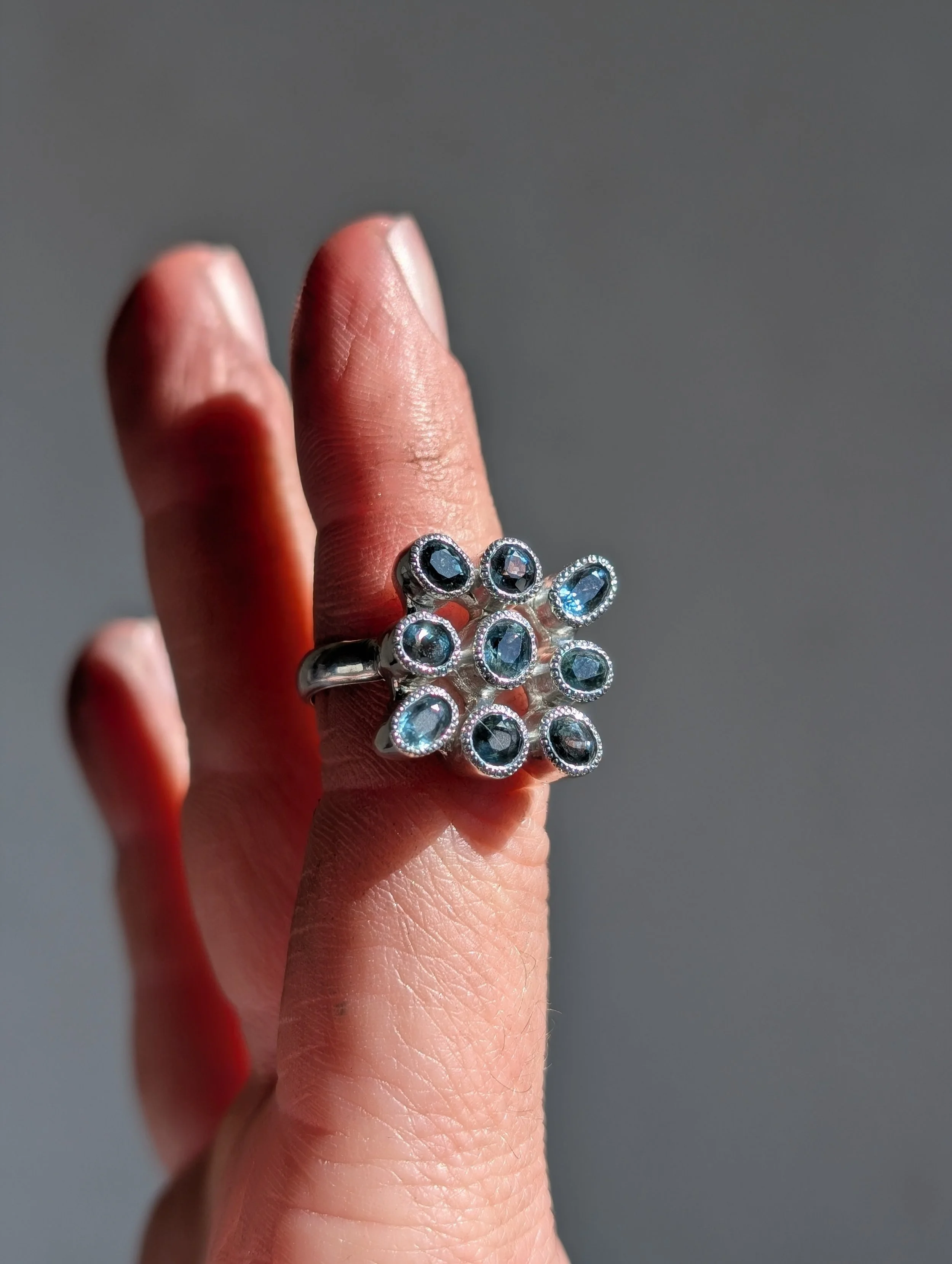A person's finger wearing a floral-shaped ring with multiple blue gemstones set in a silver band.