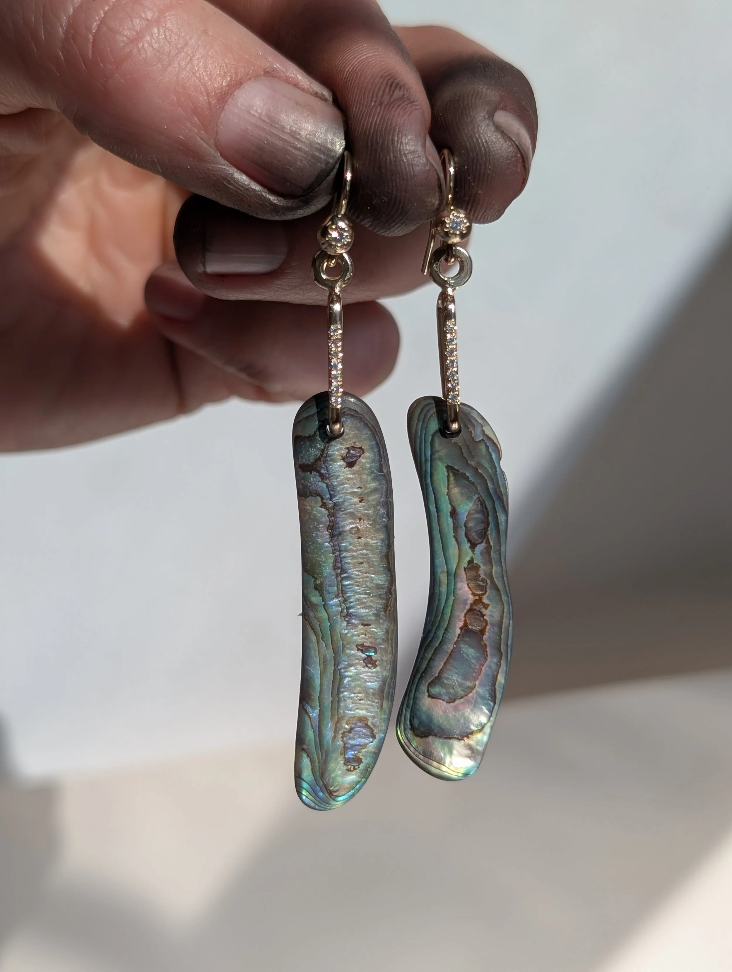 Hand holding a pair of pearl earrings with abalone shell pendants and diamond accents.