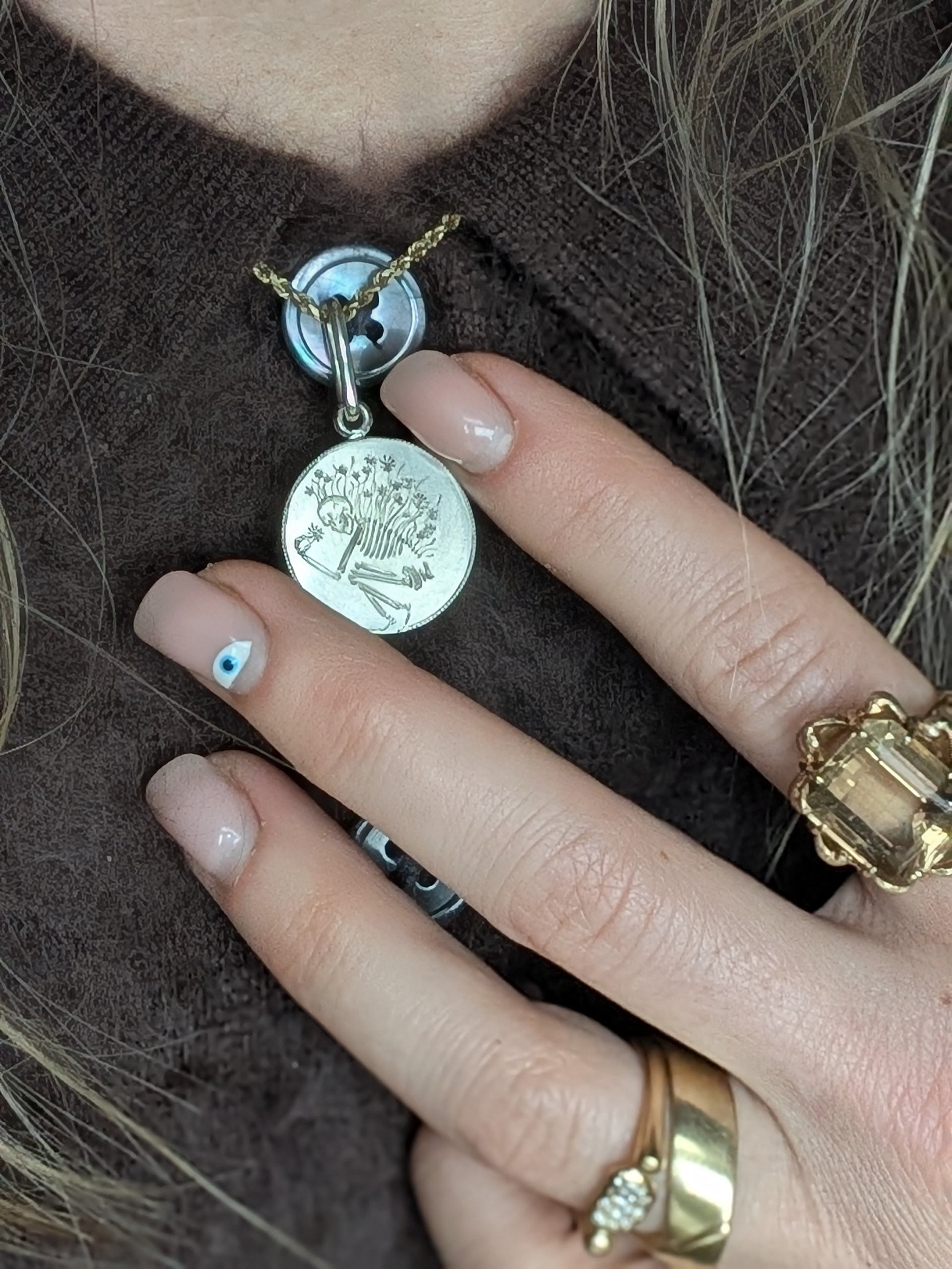 Close-up of a person's hand with rings and a painted nail, resting on a dark brown fabric, with a necklace with a coin pendant and a button visible.