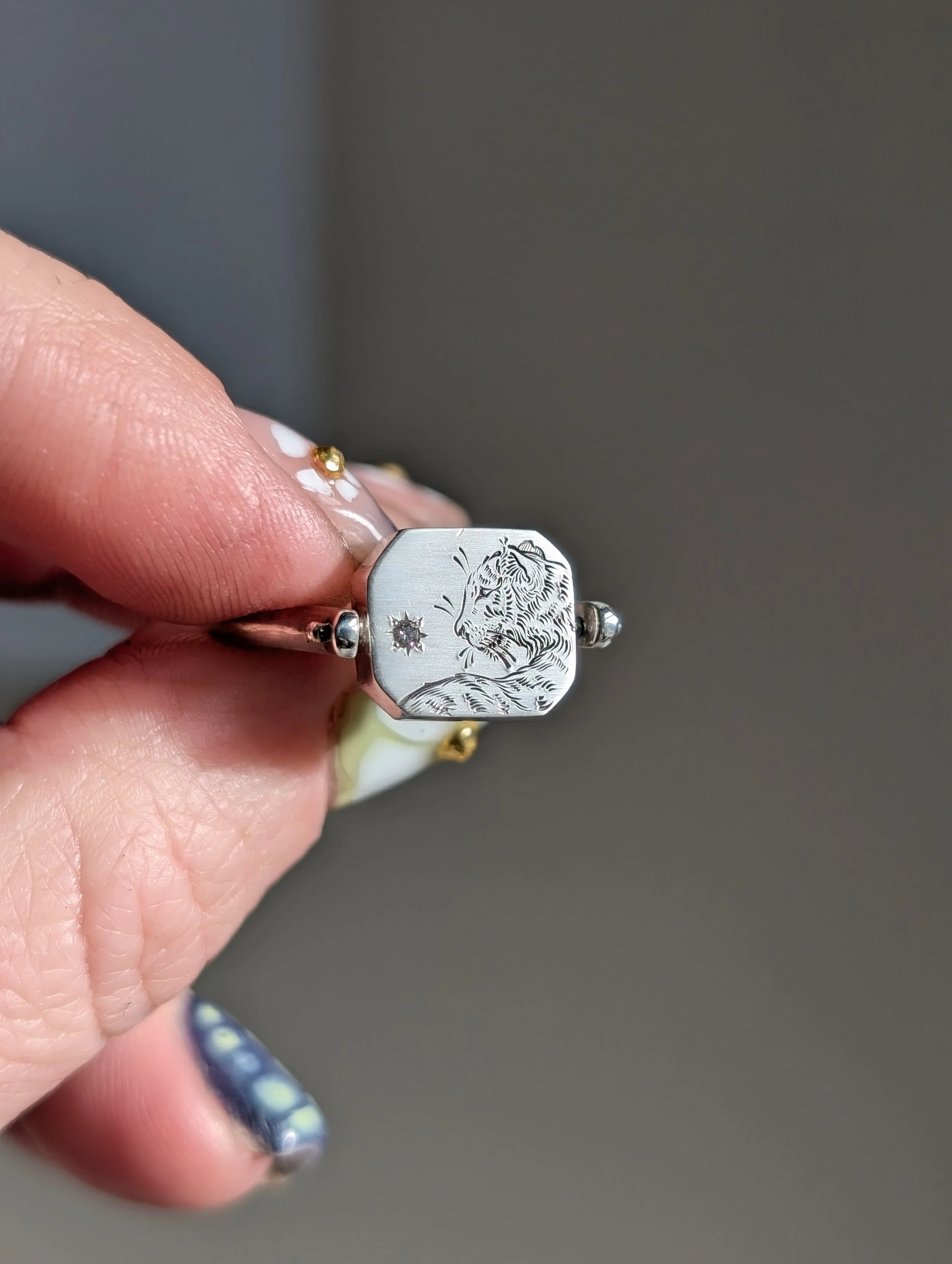 A hand holding a silver ring with an engraved tiger face and a small star-shaped gemstone, with fingernails painted with blue and yellow dots.