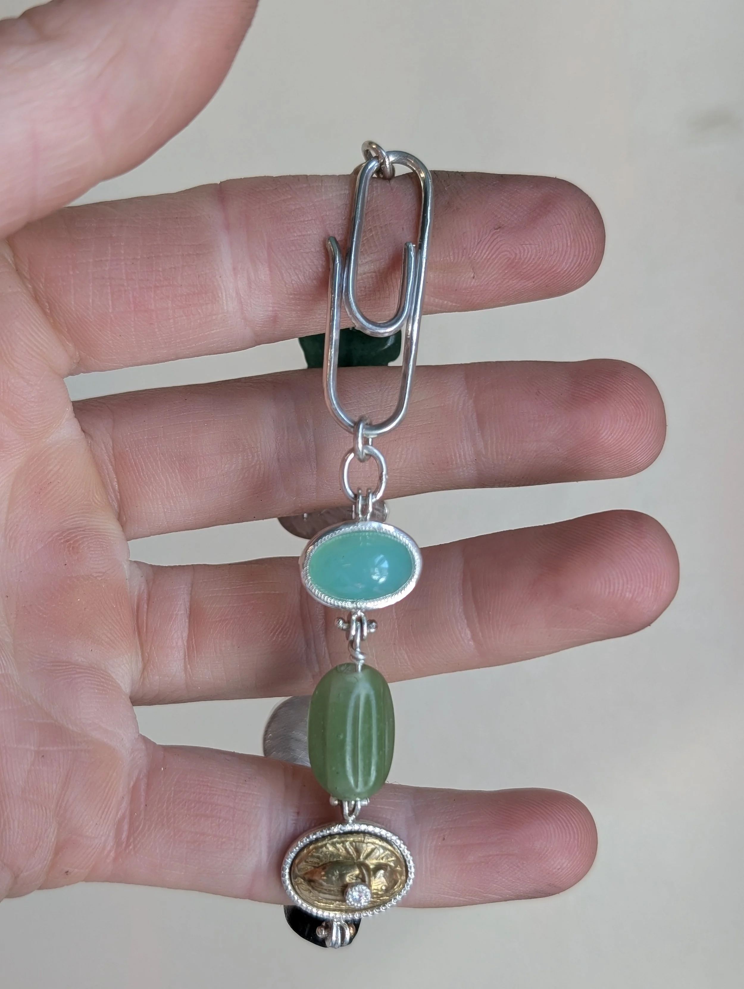 A hand holding a silver necklace with a large paperclip at the top and three charms: a round turquoise stone, a green bead, and a circular gold pendant with a diamond accent.