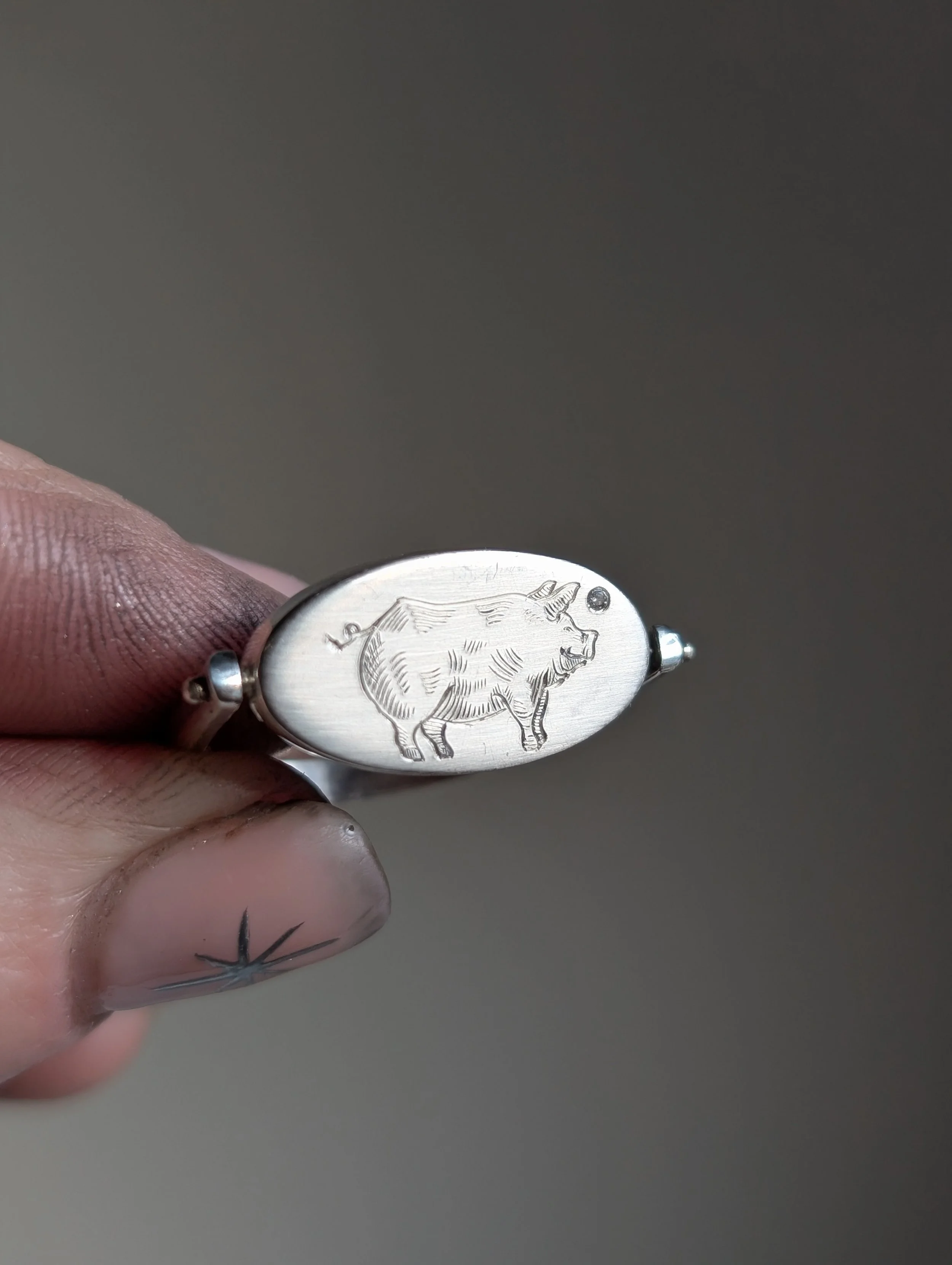 A hand holding a silver ring with a carved image of a boar on its oval face.