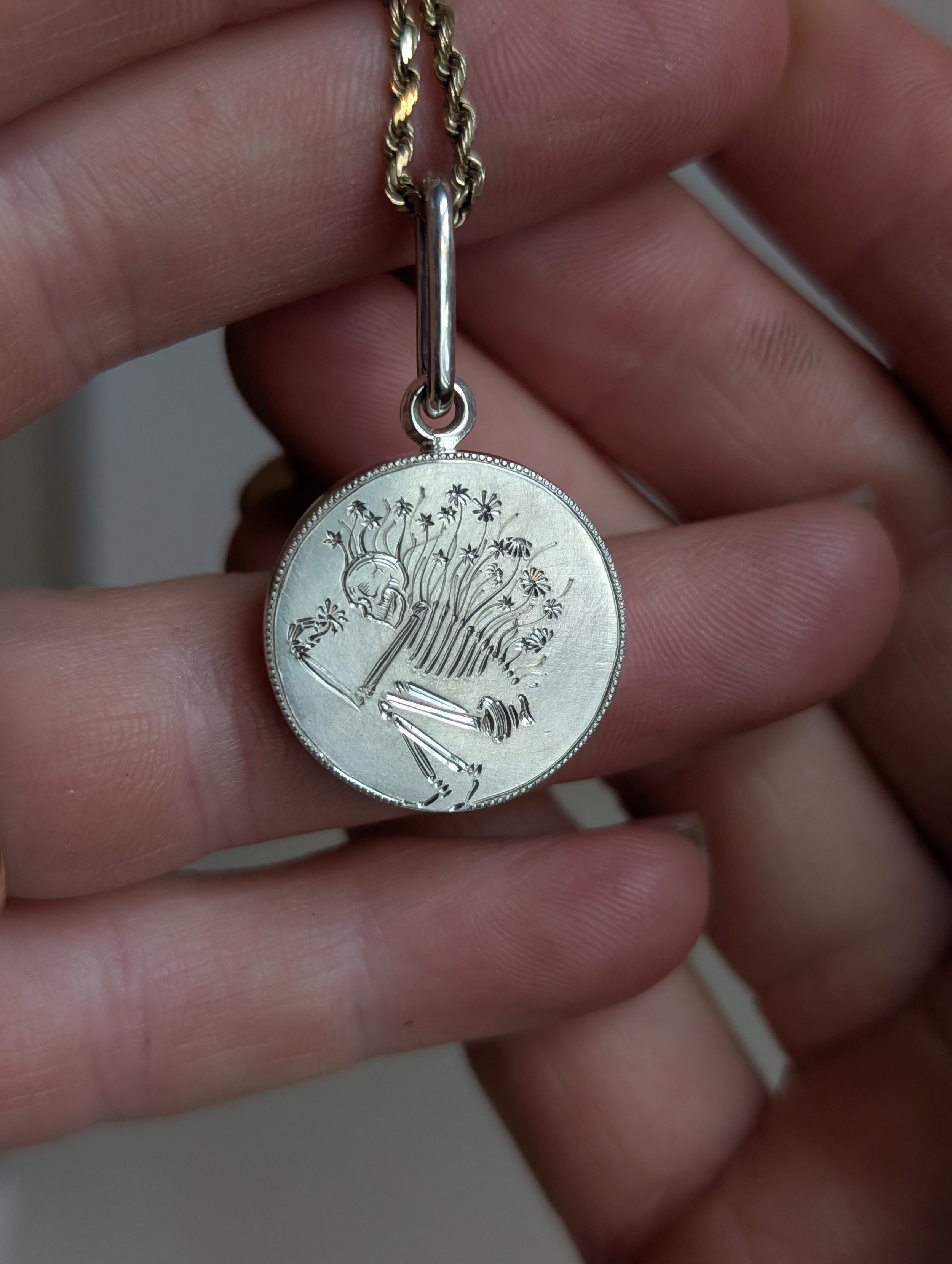 A close-up photo of a person holding a silver medal with a tree and a skull engraved on its surface, attached to a gold chain necklace.
