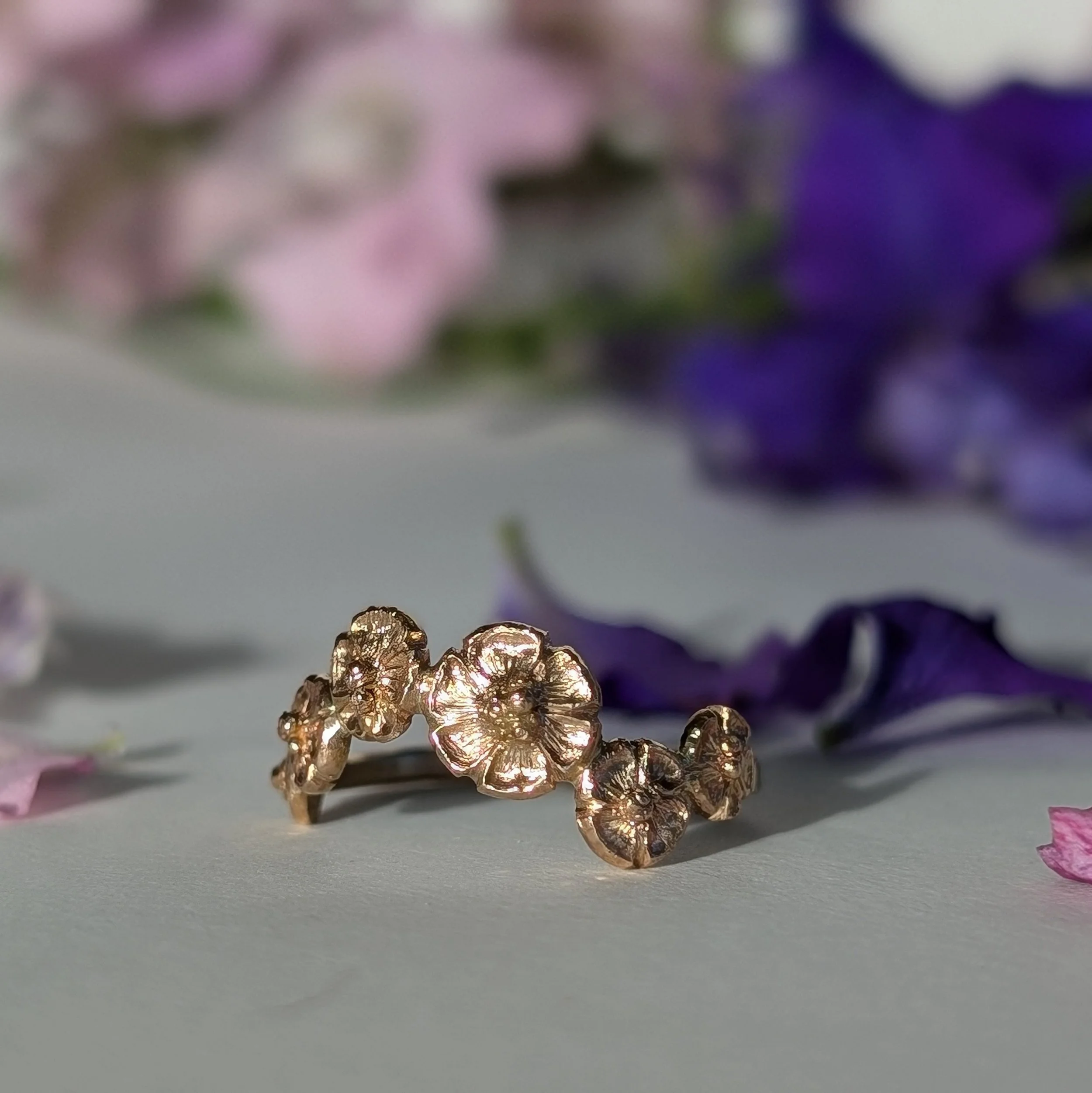 Close-up of a gold floral ring surrounded by pink and purple flower petals on a white surface.