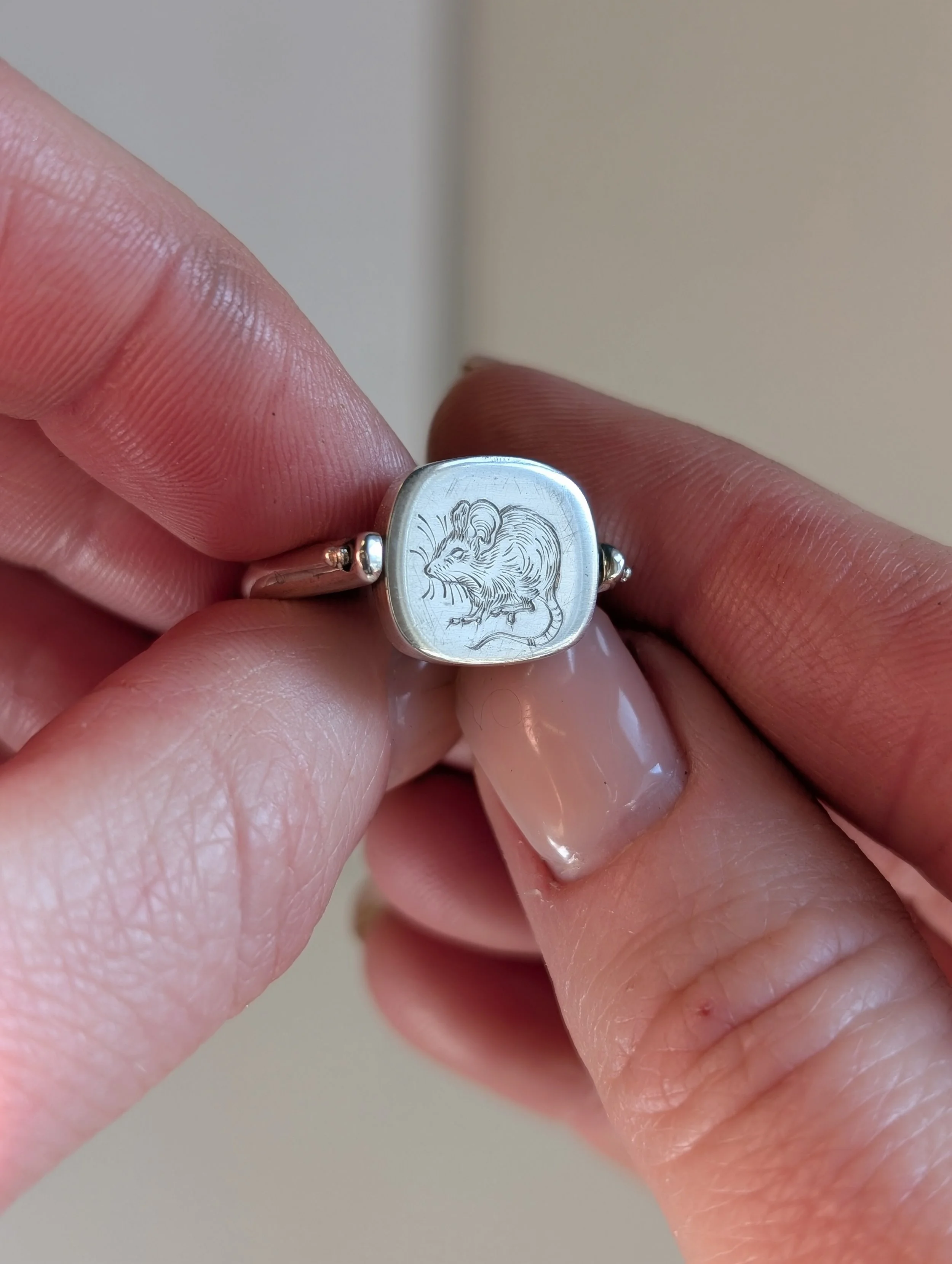 A silver ring with a square face engraved with a mouse image, held between person's thumb and index finger.