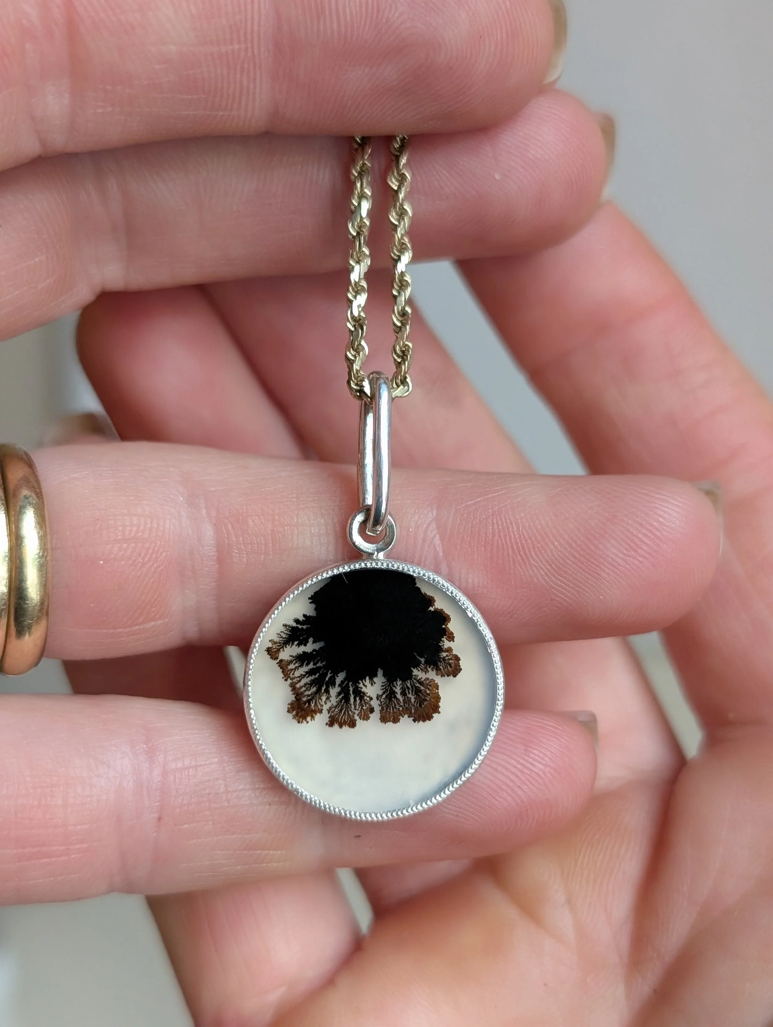 Pendant necklace with a round charm featuring a black and brown fern leaf design on a white background, held by a person with gold jewelry.