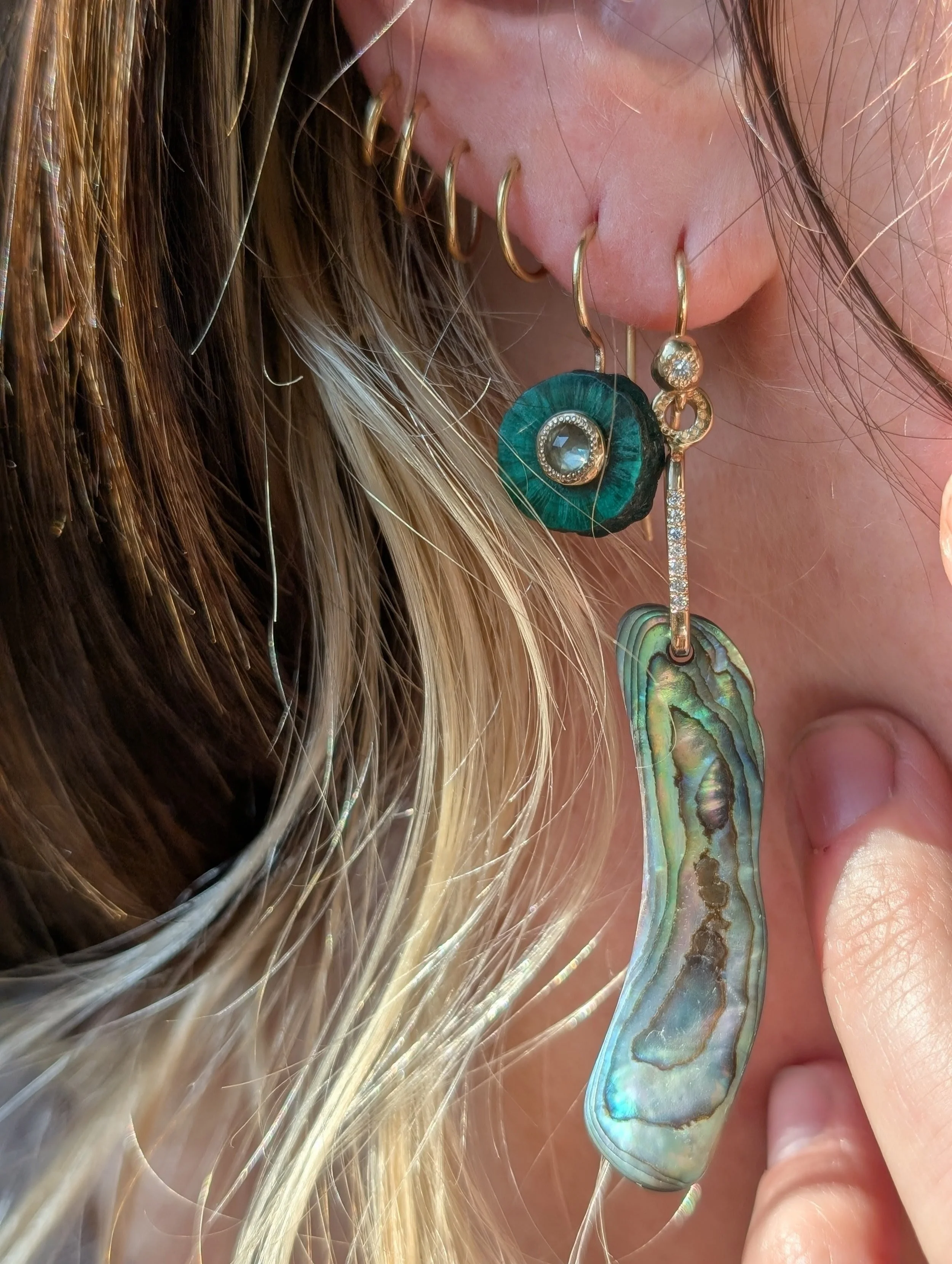 Close-up of a woman's face showing multiple gold earrings, including a large, curved, iridescent shell or mother-of-pearl earring, and a dark green circular earring with a central gemstone, with her blond hair partially covering her face.