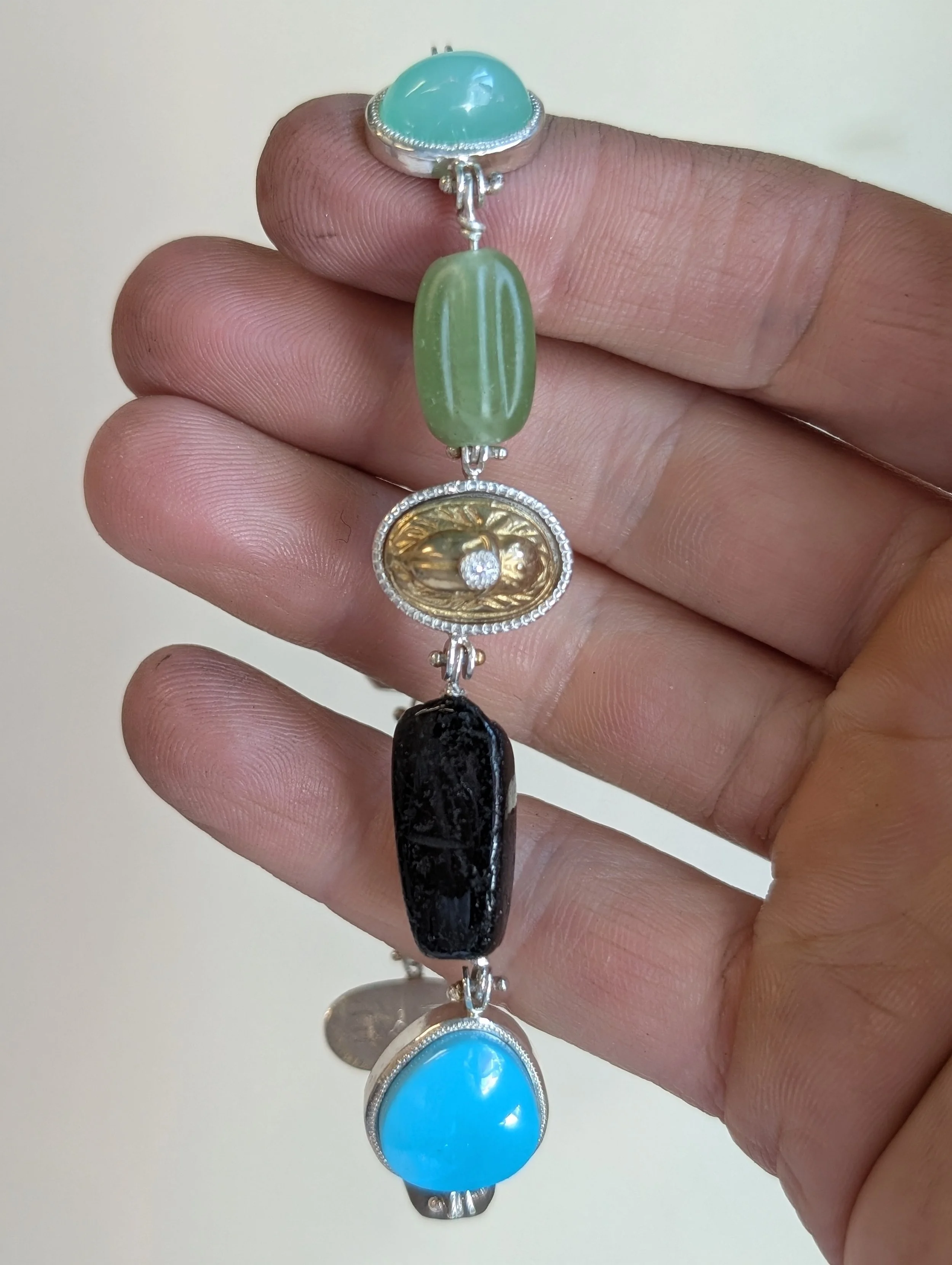Hand holding a colorful multistone bracelet with blue, green, gold, black stones, and a small diamond detail.