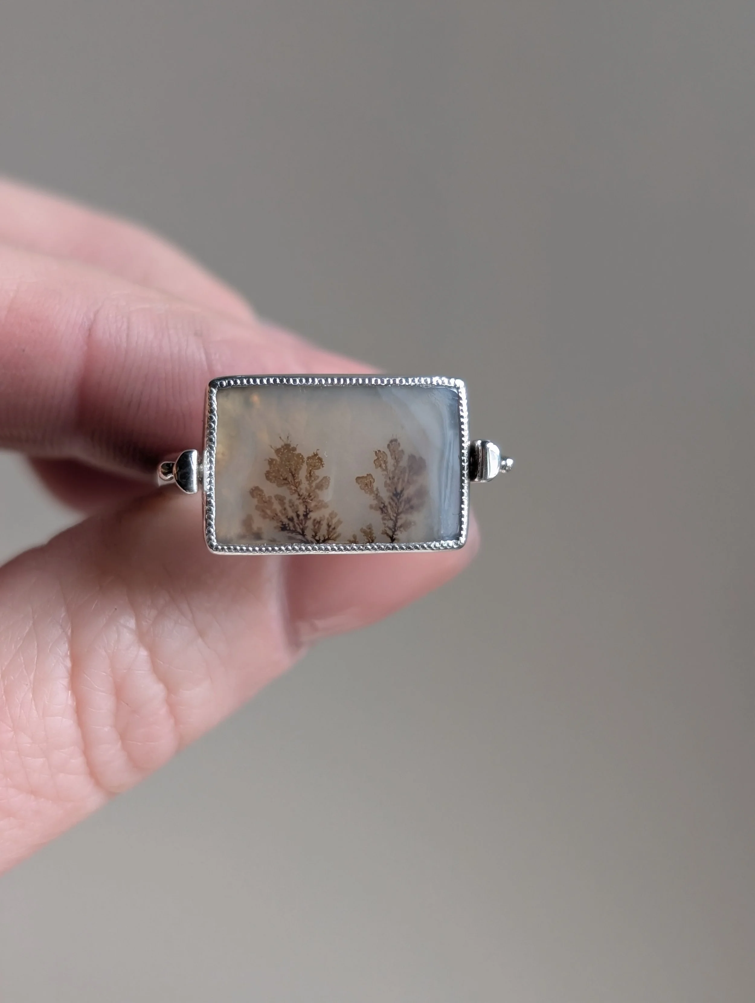 A hand holding a silver ring with a rectangular center featuring a leaf pattern encased in clear resin.