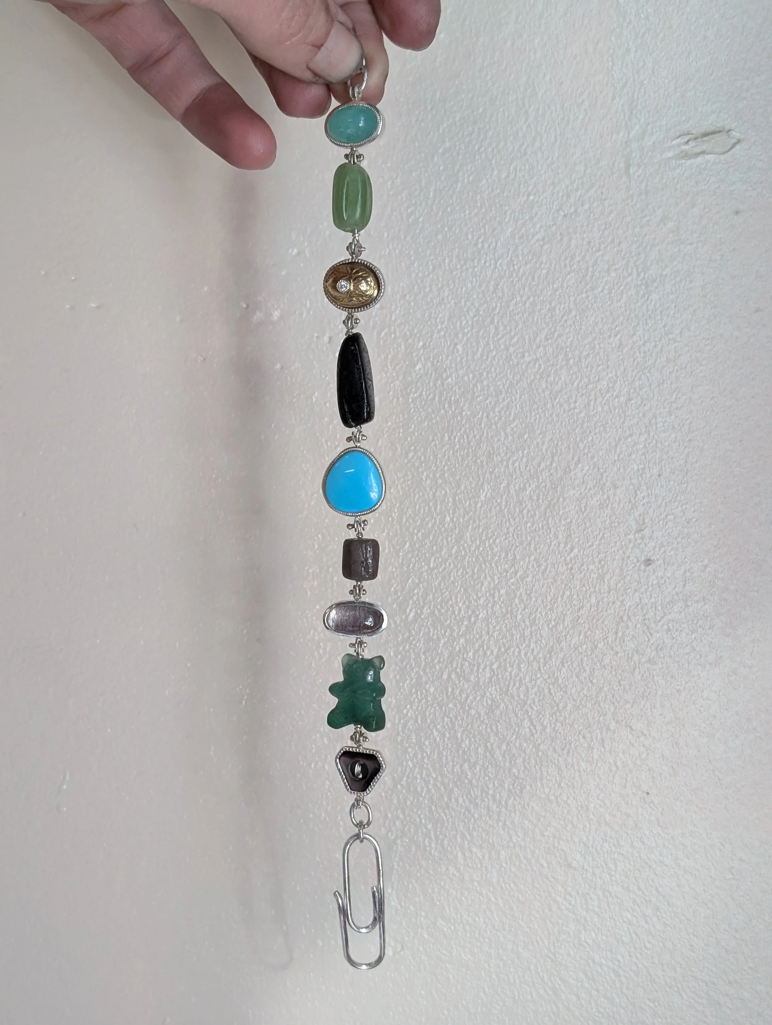 Hand holding a silver keychain with multiple colorful beads and charms, including a blue stone, a green bead, and a teddy bear-shaped charm, against a white wall.