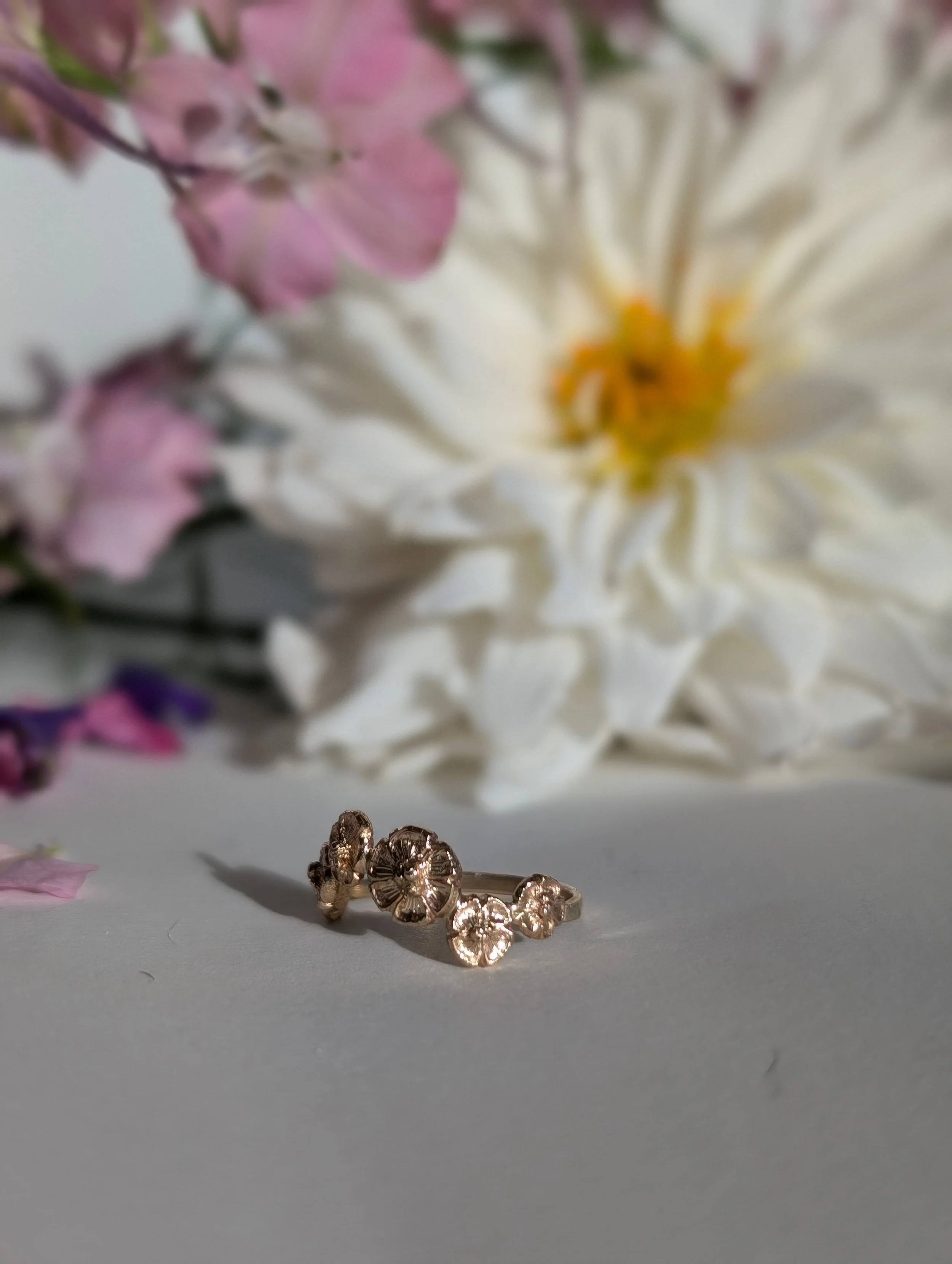 Close-up of a gold ring with floral design on a white surface, with blurred pink and white flowers in the background.