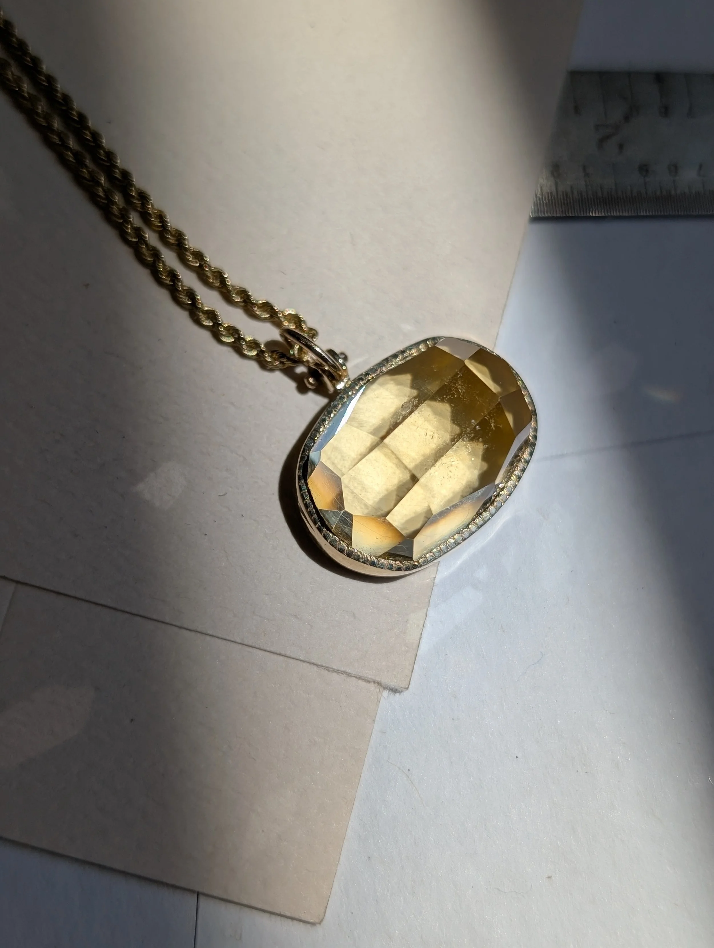 A gold necklace with a large, oval, faceted yellow gemstone pendant, surrounded by small clear stones, on white paper.