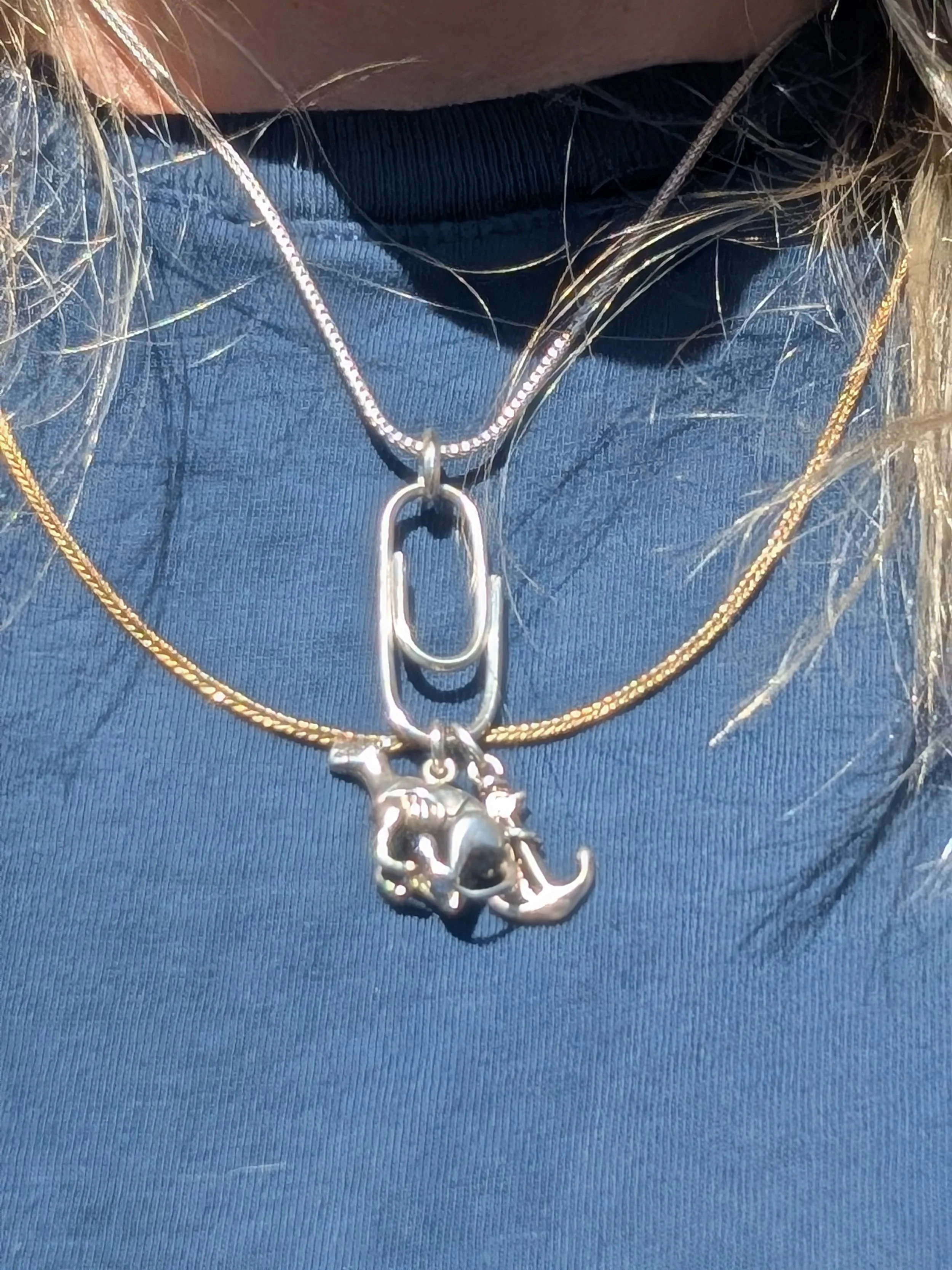 Close-up of a person's neck wearing layered necklaces, including a silver chain with a lock-shaped pendant, a gold chain, and a silver chain with a charm resembling a whale and a crescent moon, on a dark blue shirt.
