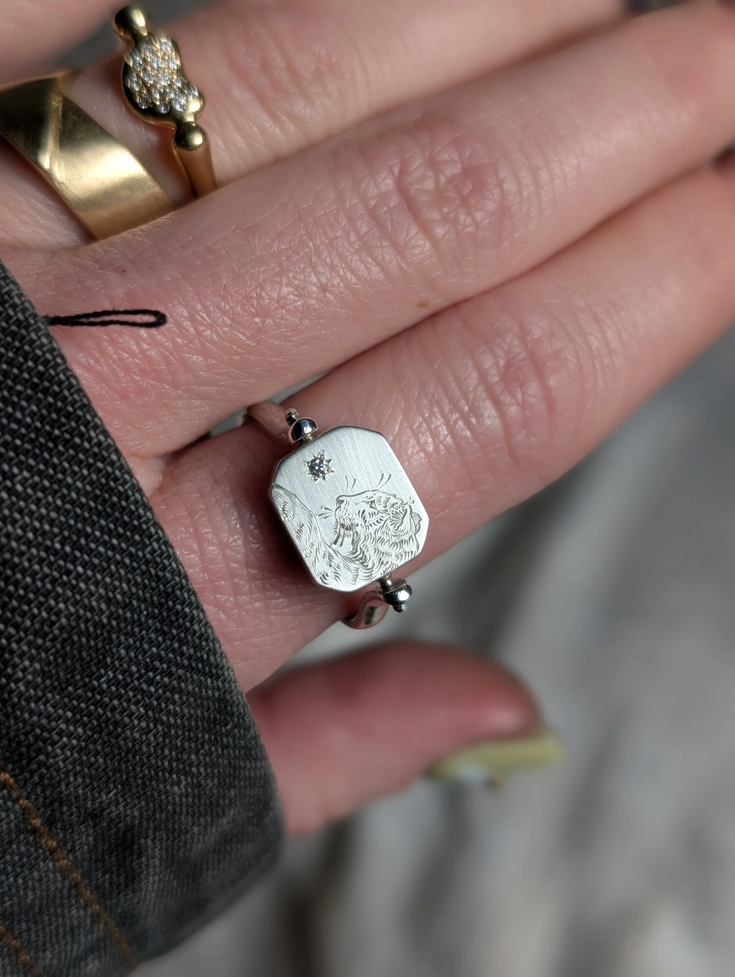 Close-up of a hand with two rings, one gold and one silver. The silver ring features a lion's head engraving and a small star-shaped gemstone. The person is wearing a dark, textured garment and is holding their hand in a relaxed position.
