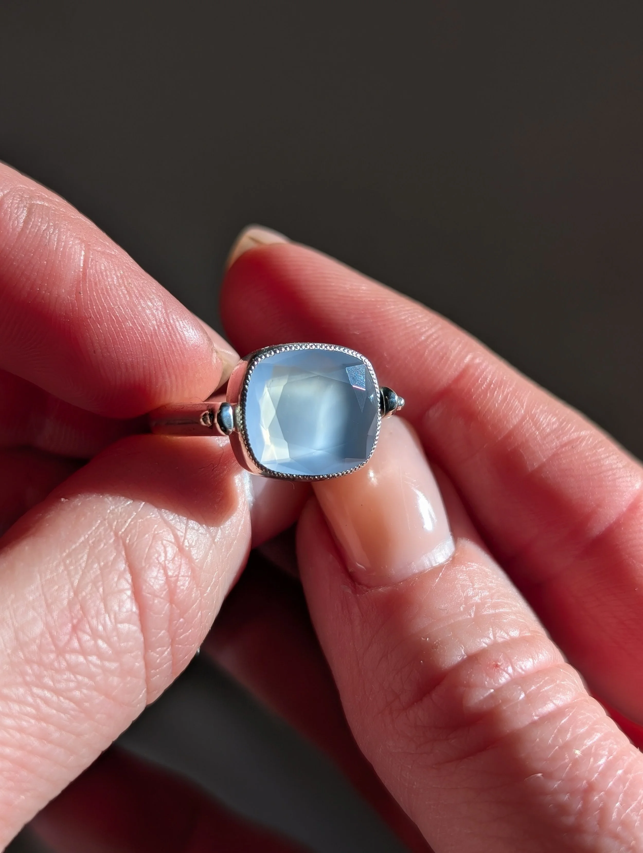 Close-up of a hand holding a silver ring with a large, faceted, square-shaped, light blue gemstone.