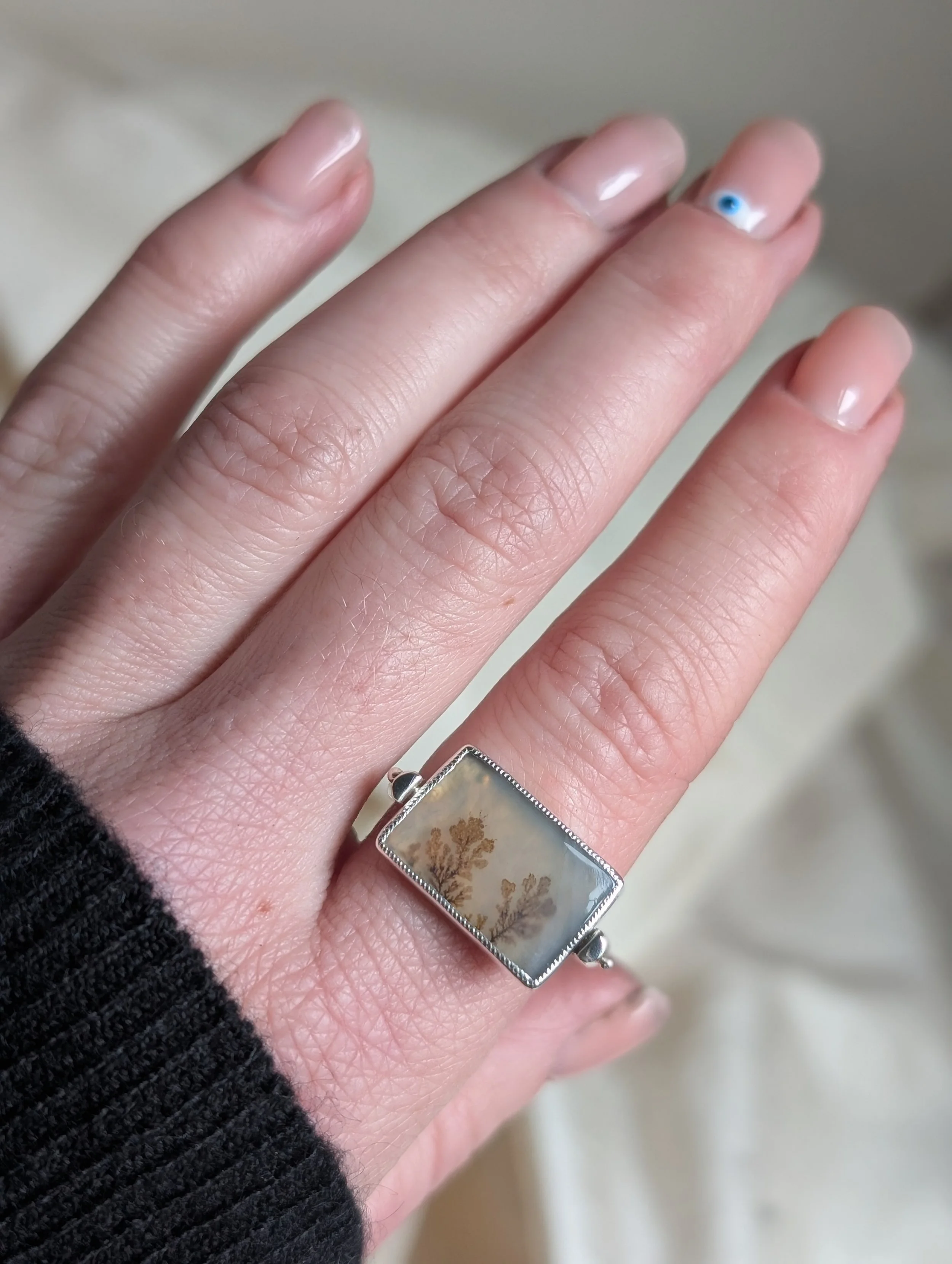 Close-up of a hand wearing a silver ring with a rectangular stone featuring dried botanical designs, with neatly manicured nails.