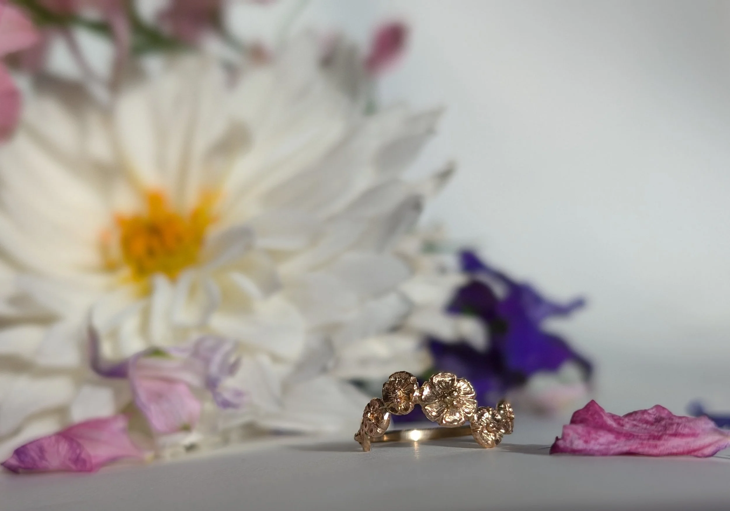 A gold ring with floral design and textured surface surrounded by white, purple, and pink flower petals, with larger flowers blurred in the background.