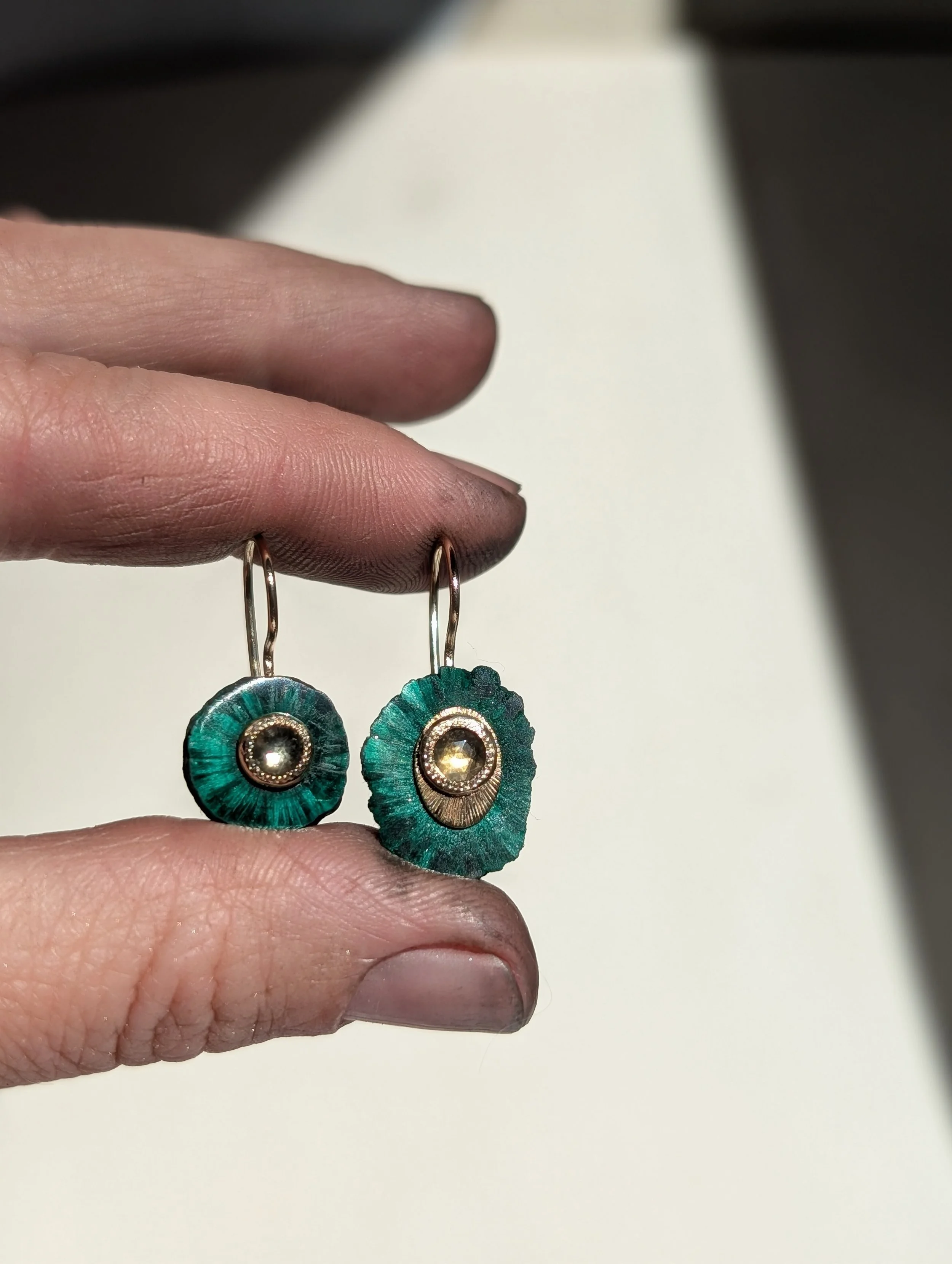 Pair of earrings with circular green and gold design, featuring a central gemstone, held by a person's hand.