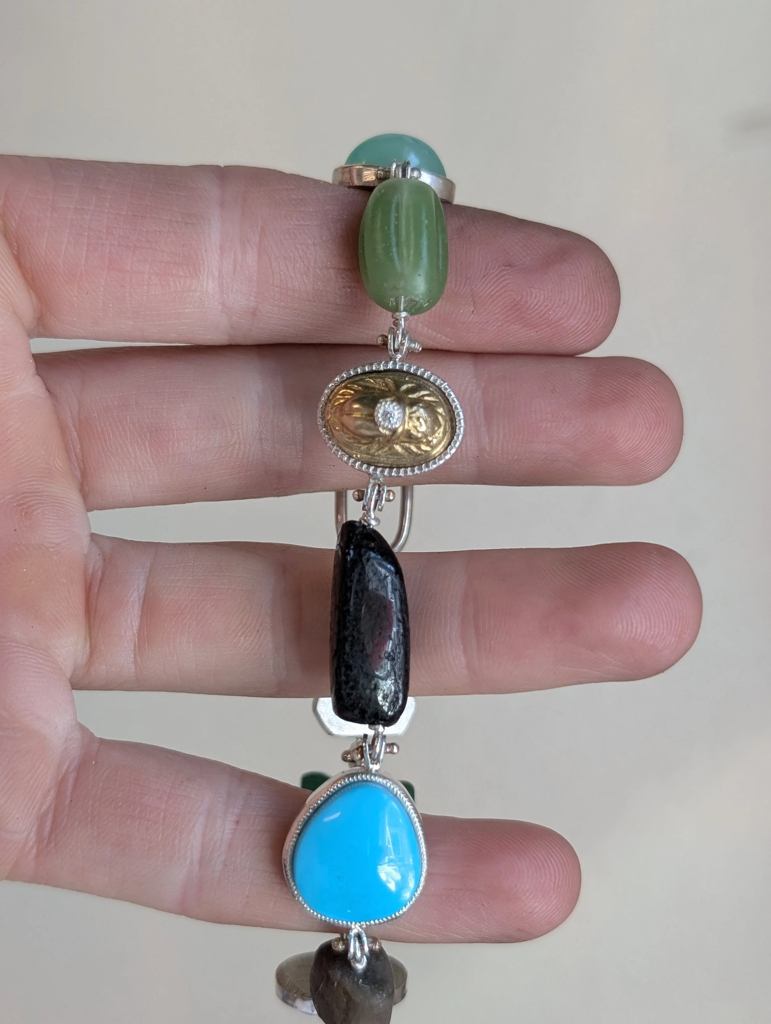 A hand holding a bracelet with five large, uniquely shaped stones in different colors and a small decorative gold face pendant with a diamond in the center.