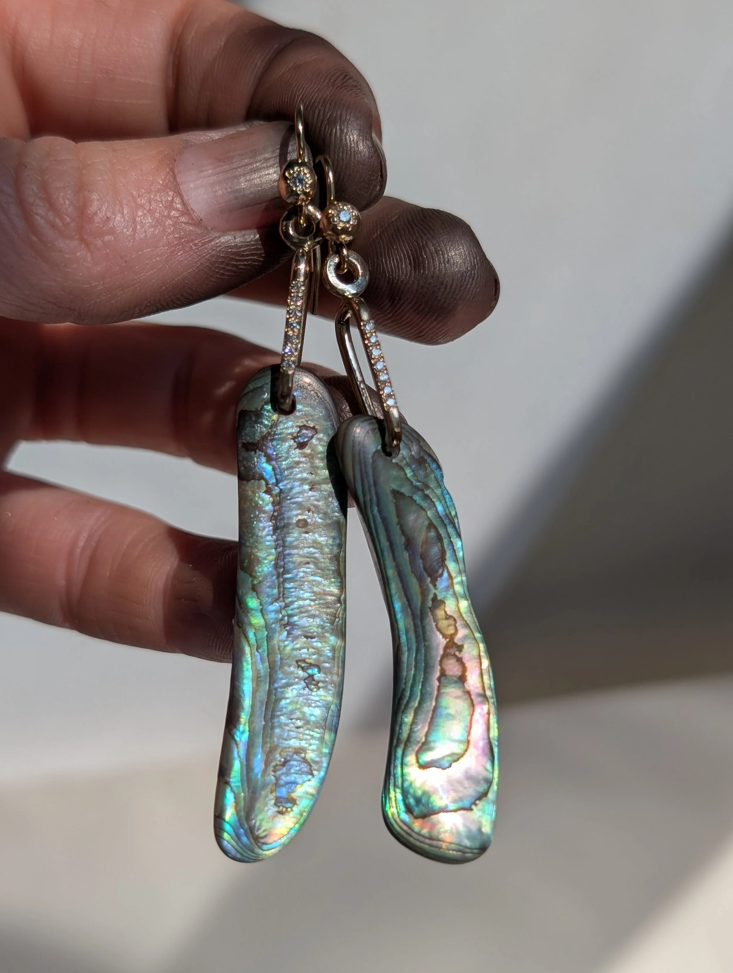 Close-up of a hand holding a pair of earrings featuring abalone shell pendants with iridescent colors, attached to gold hooks adorned with small diamonds.