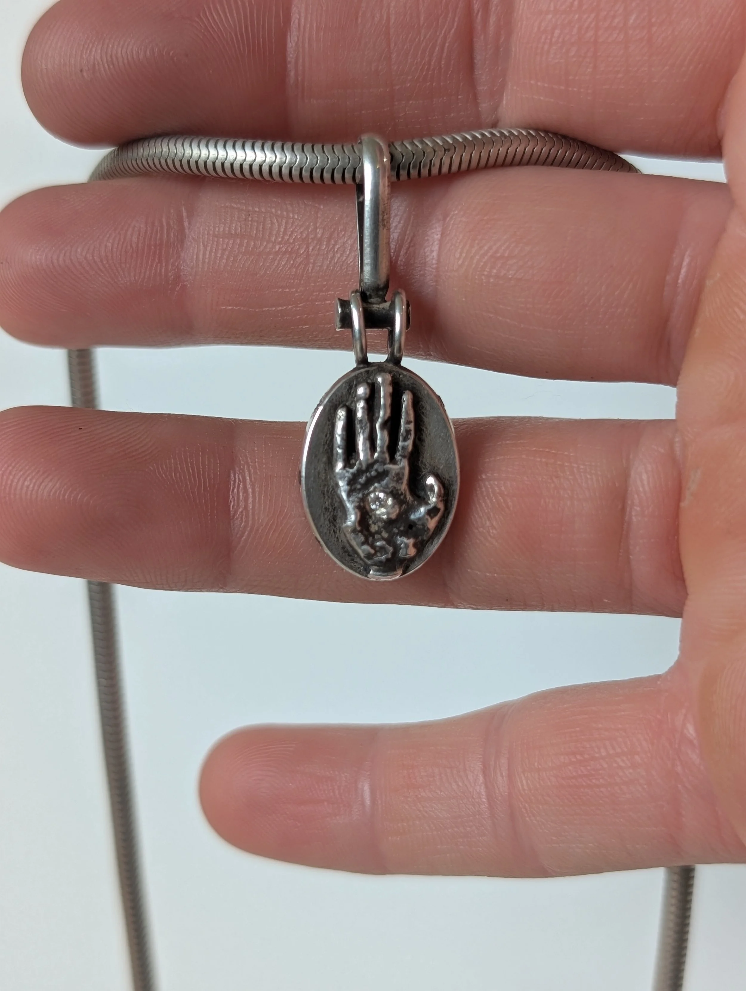 A silver bracelet with a dangling oval-shaped pendant featuring a raised handprint and a small circular design inside the palm.