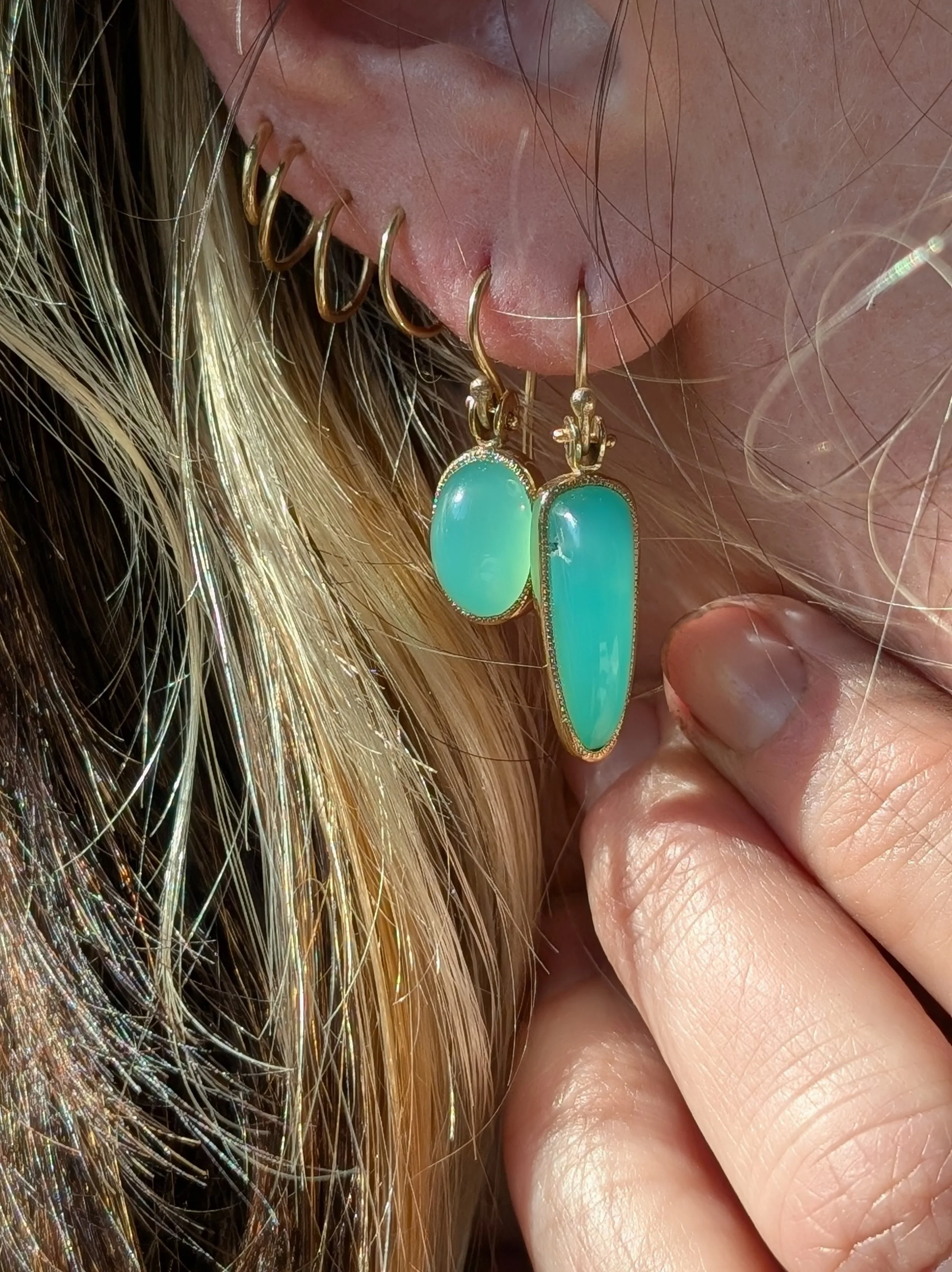 Close-up of a woman's ear with multiple gold hoop earrings, including two large turquoise-colored gemstone dangle earrings, and her hand holding her earlobe.