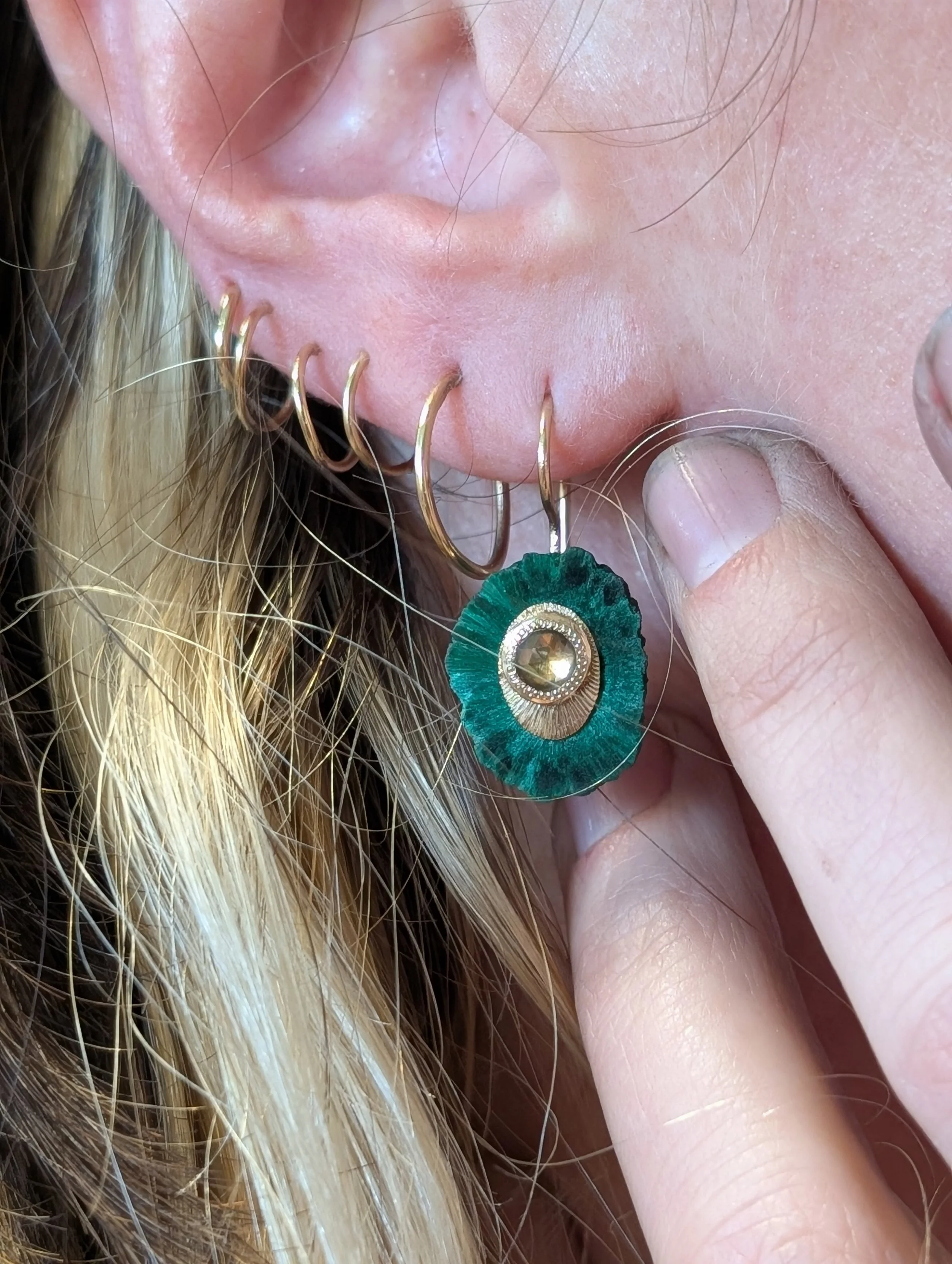 Close-up of a person with blonde hair and multiple gold hoop earrings, holding a green and gold decorative earring with a clear gemstone, and slightly touching the earring to their ear.