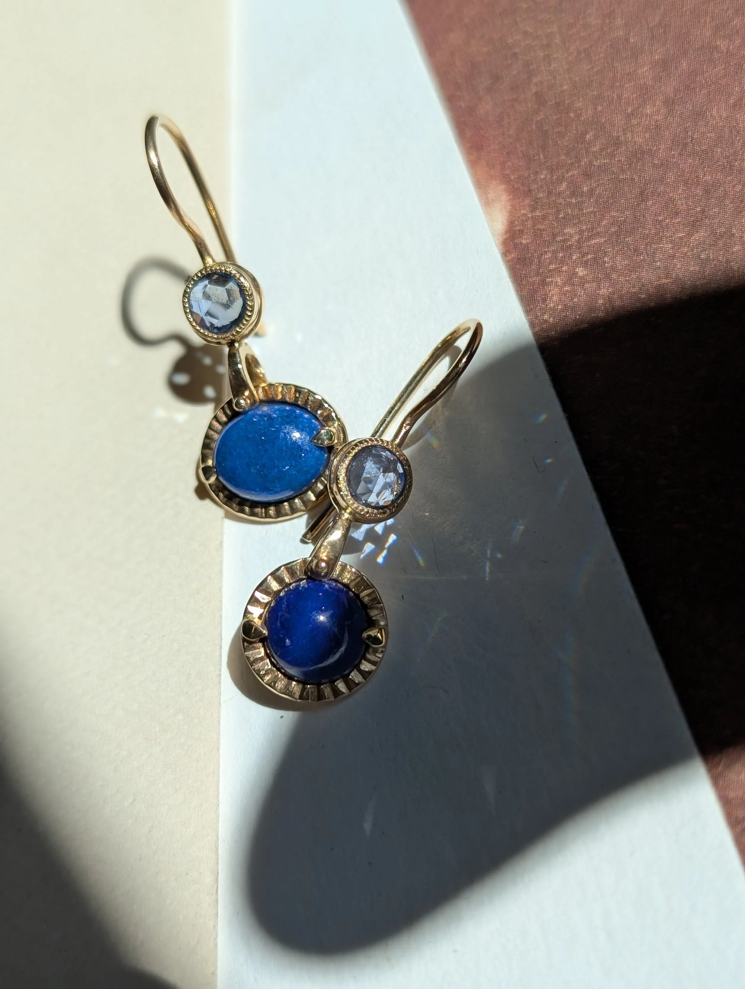 Gold earrings with blue stone accents and clear gemstones, casting shadows on a white and pink background.