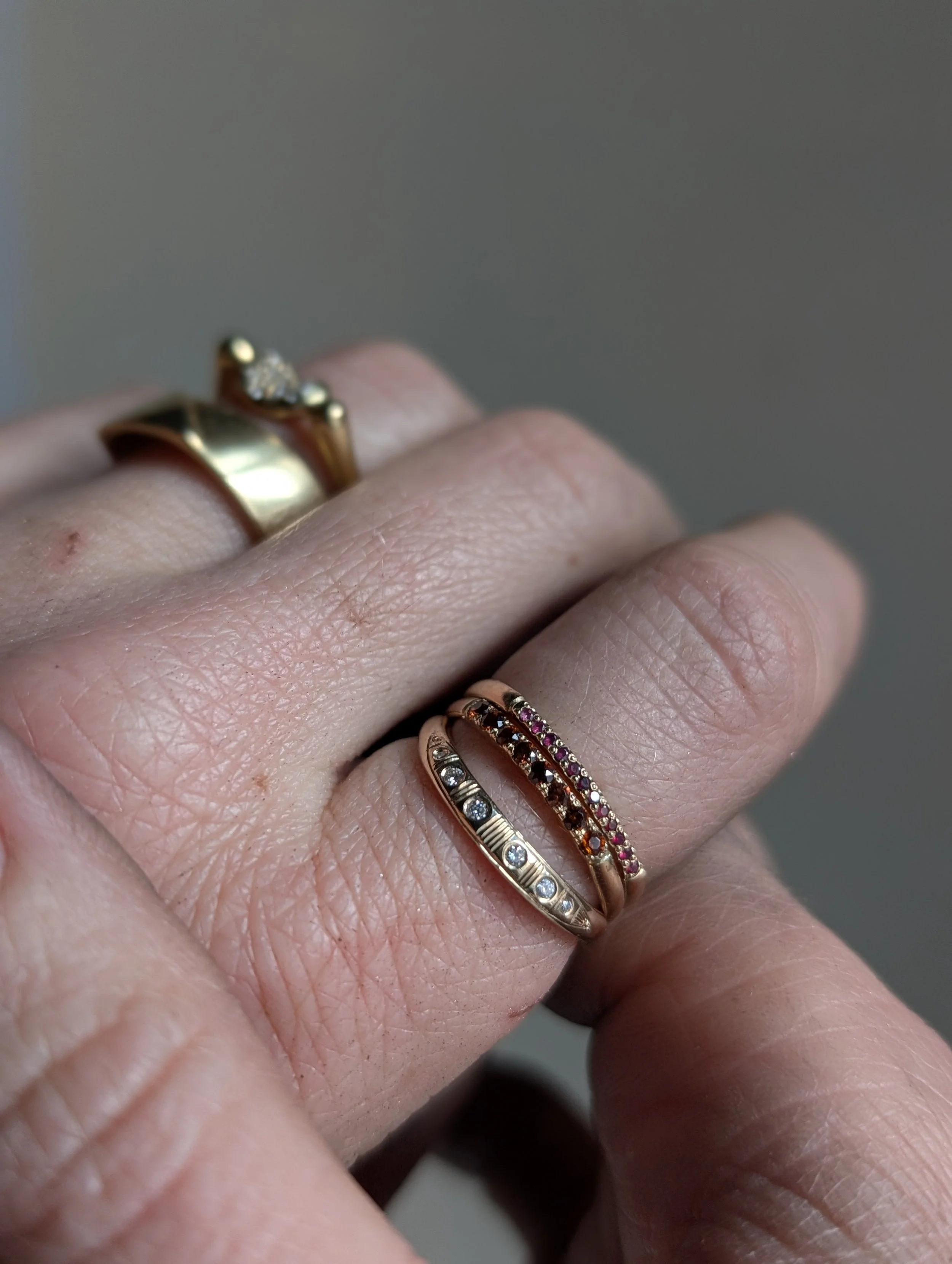Close-up of a hand wearing three rings, including a gold band and two rings with colored stones, on a light skin tone hand with visible skin texture and wrinkles.