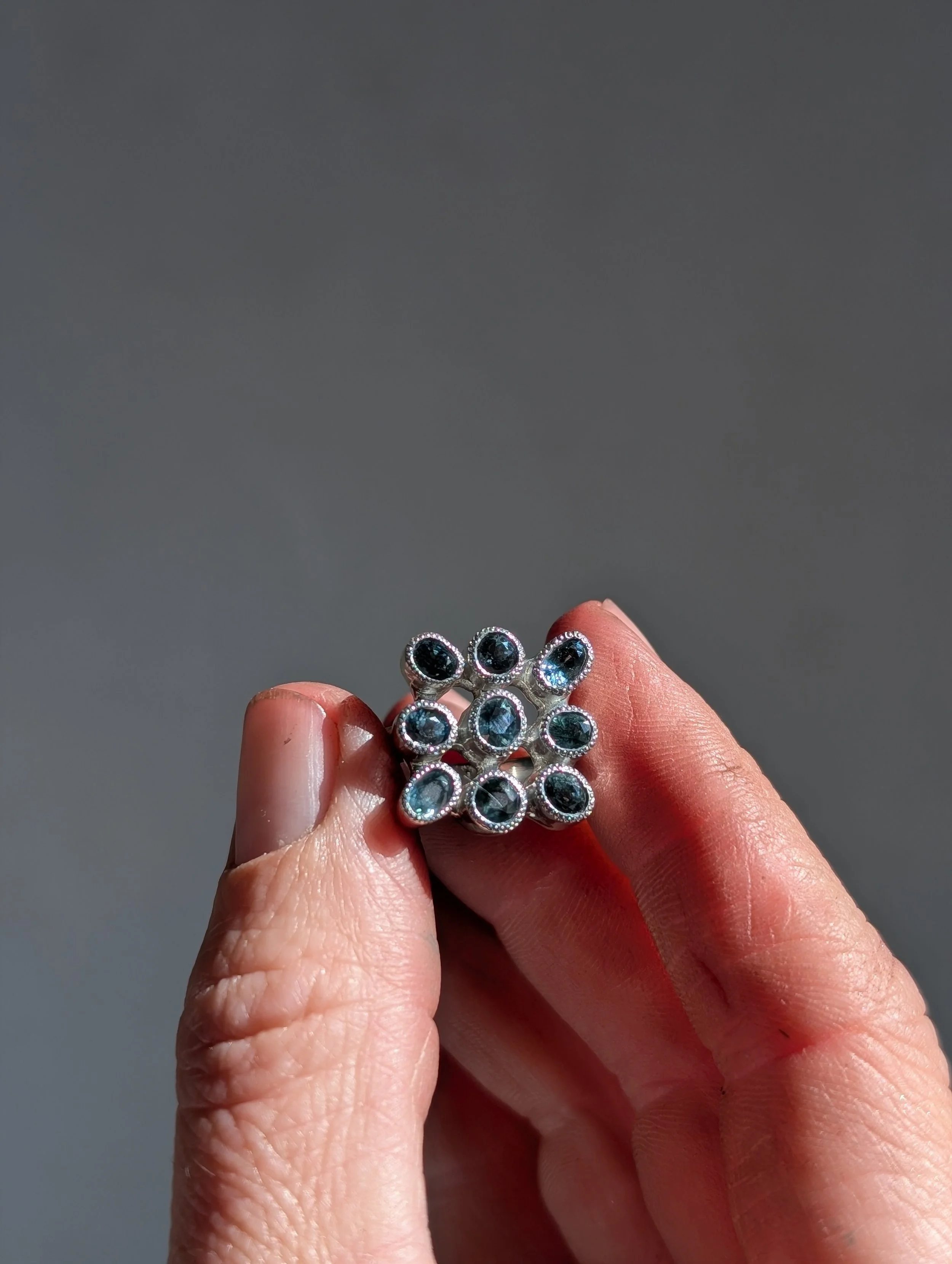 Close-up of a hand holding a silver ring with multiple black and blue stones arranged in a circular pattern.