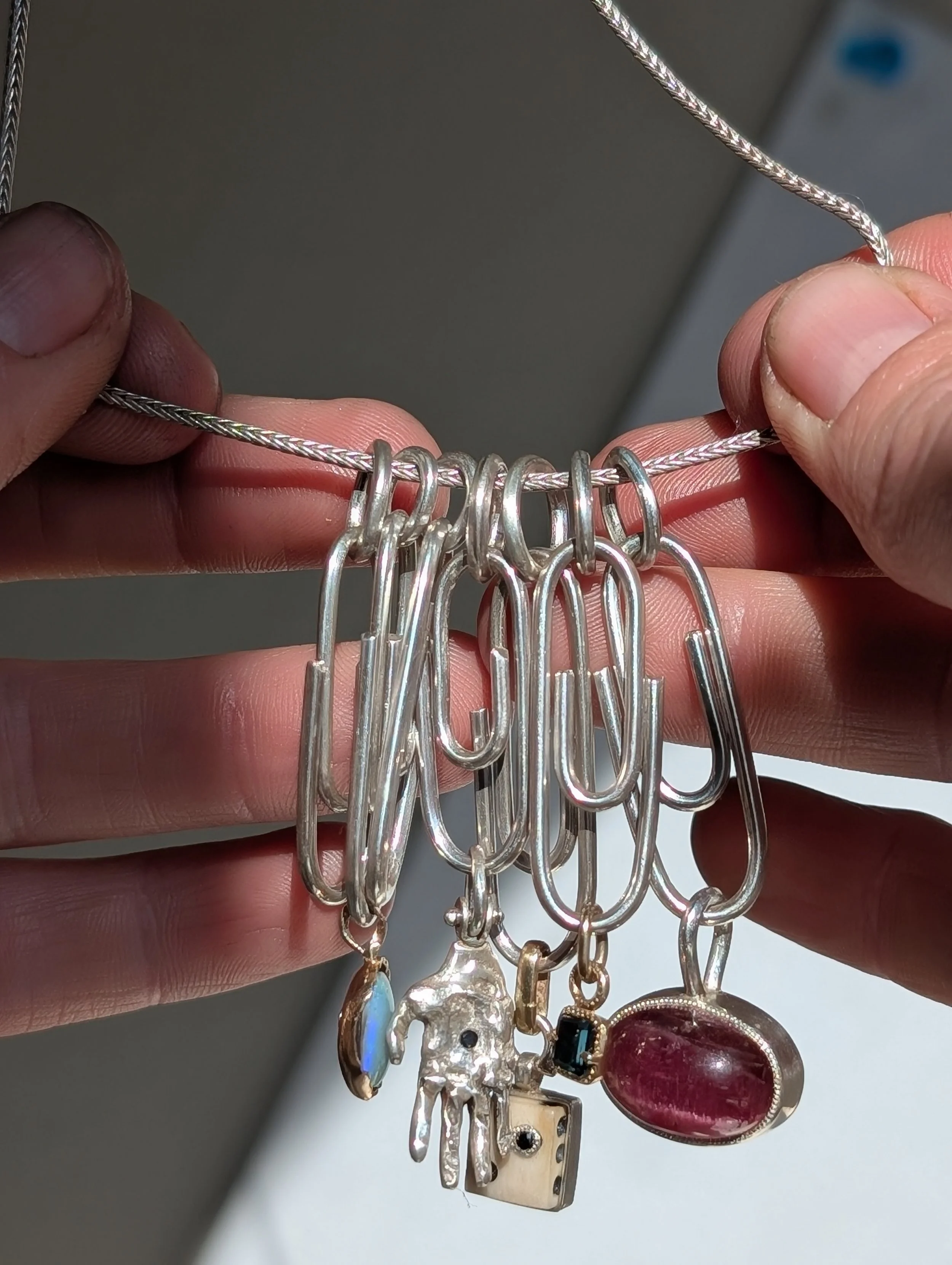 A person's hands holding a silver necklace with multiple large, elongated safety pins attached, some with small charms including a hand, a gemstone, and a lock.