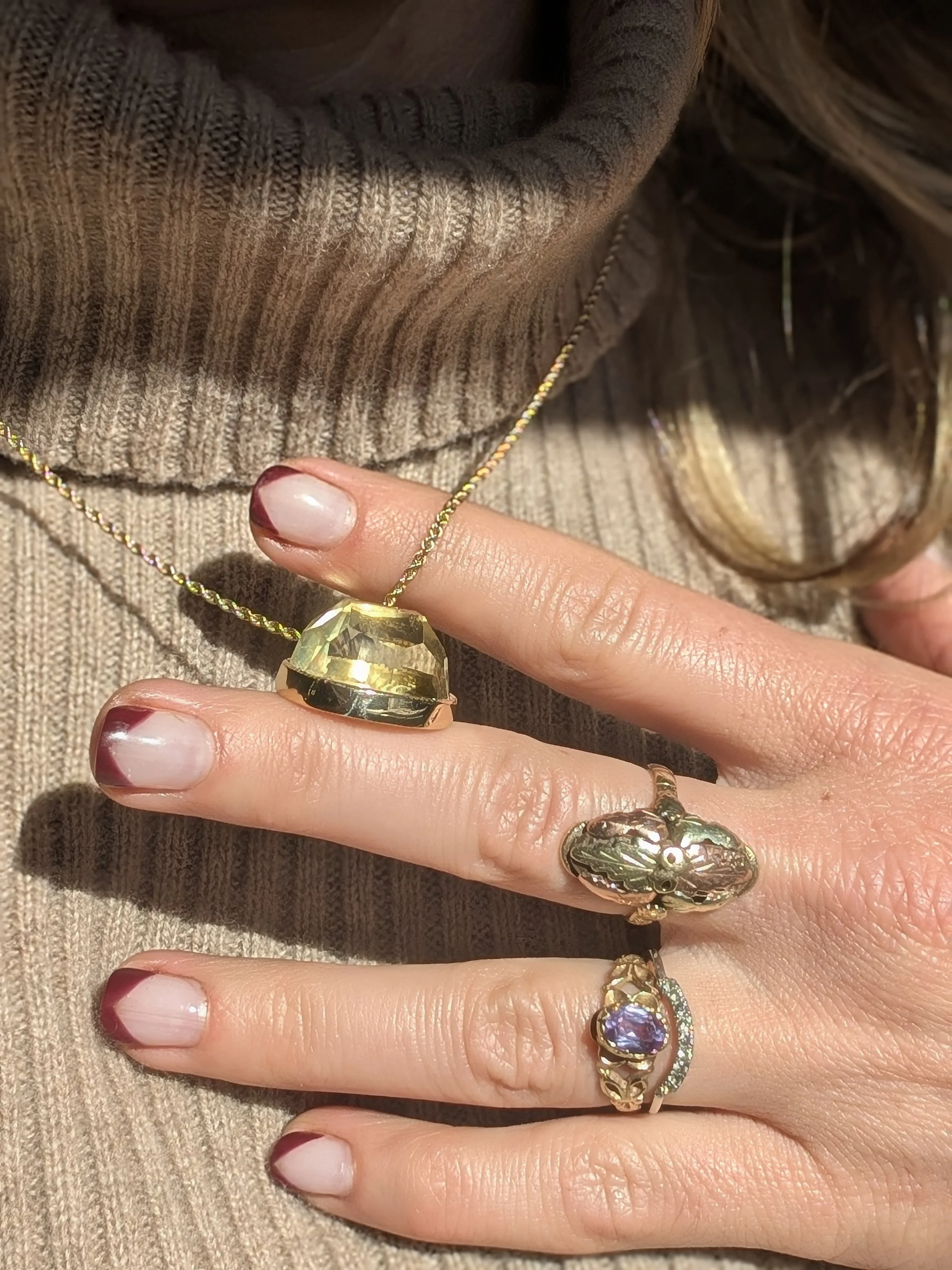 Close-up of a woman's hand wearing multiple rings, holding a gold pendant necklace with a large faceted gemstone. The woman is wearing a beige ribbed sweater.